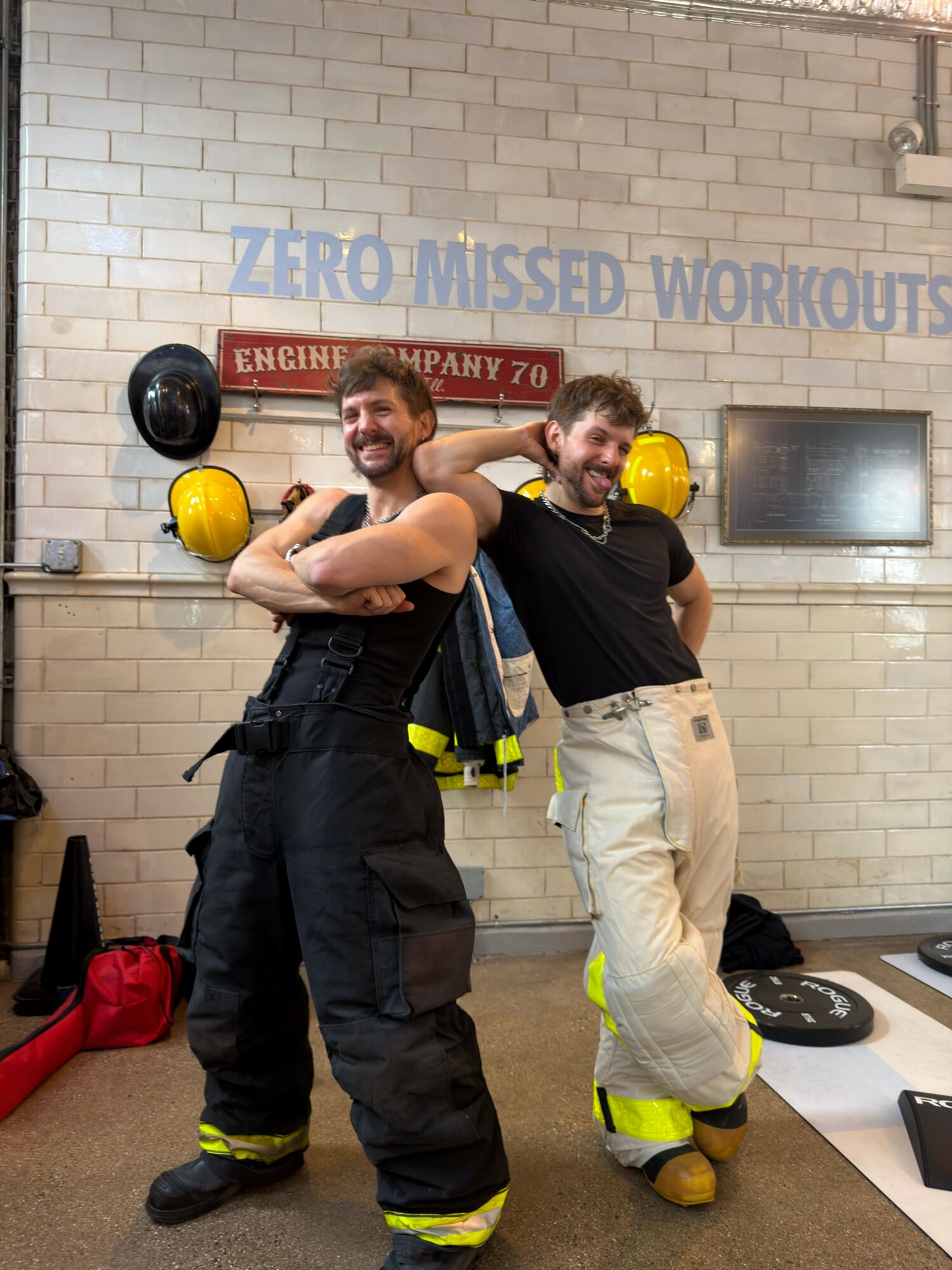 Two firefighters in uniform pose with helmets in a fire station, smiling and flexing muscles, with equipment and a brick wall background.