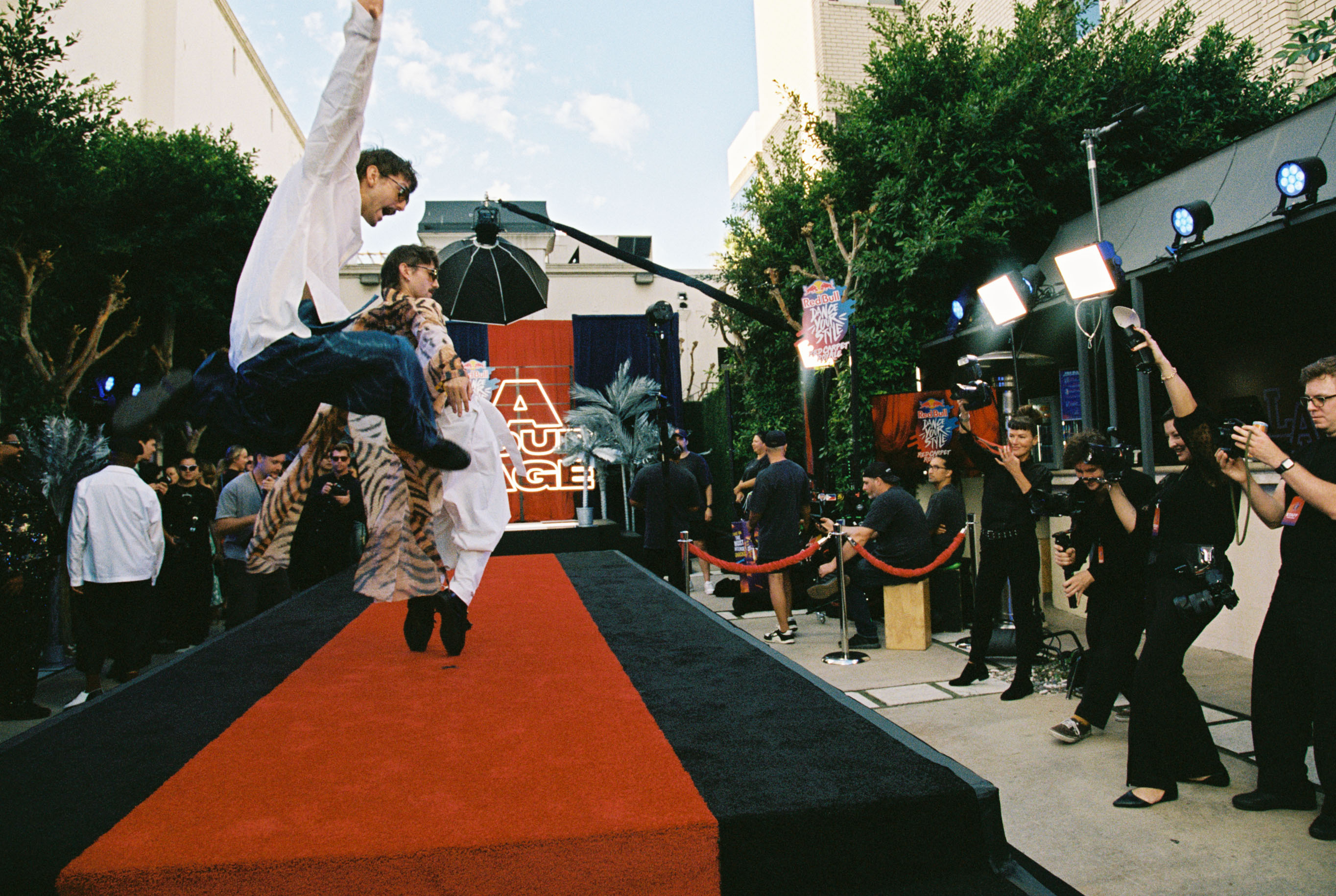 A performer jumps on a red carpet runway during an outdoor event with a crowd and stage lighting.