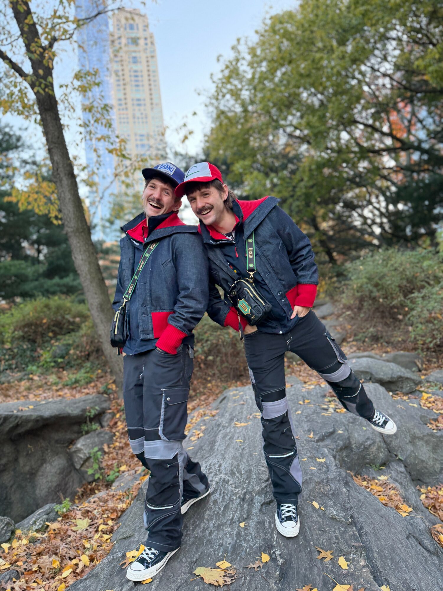 Two people in outdoor clothing standing on a rock in a park with trees and a city skyline in the background, smiling.