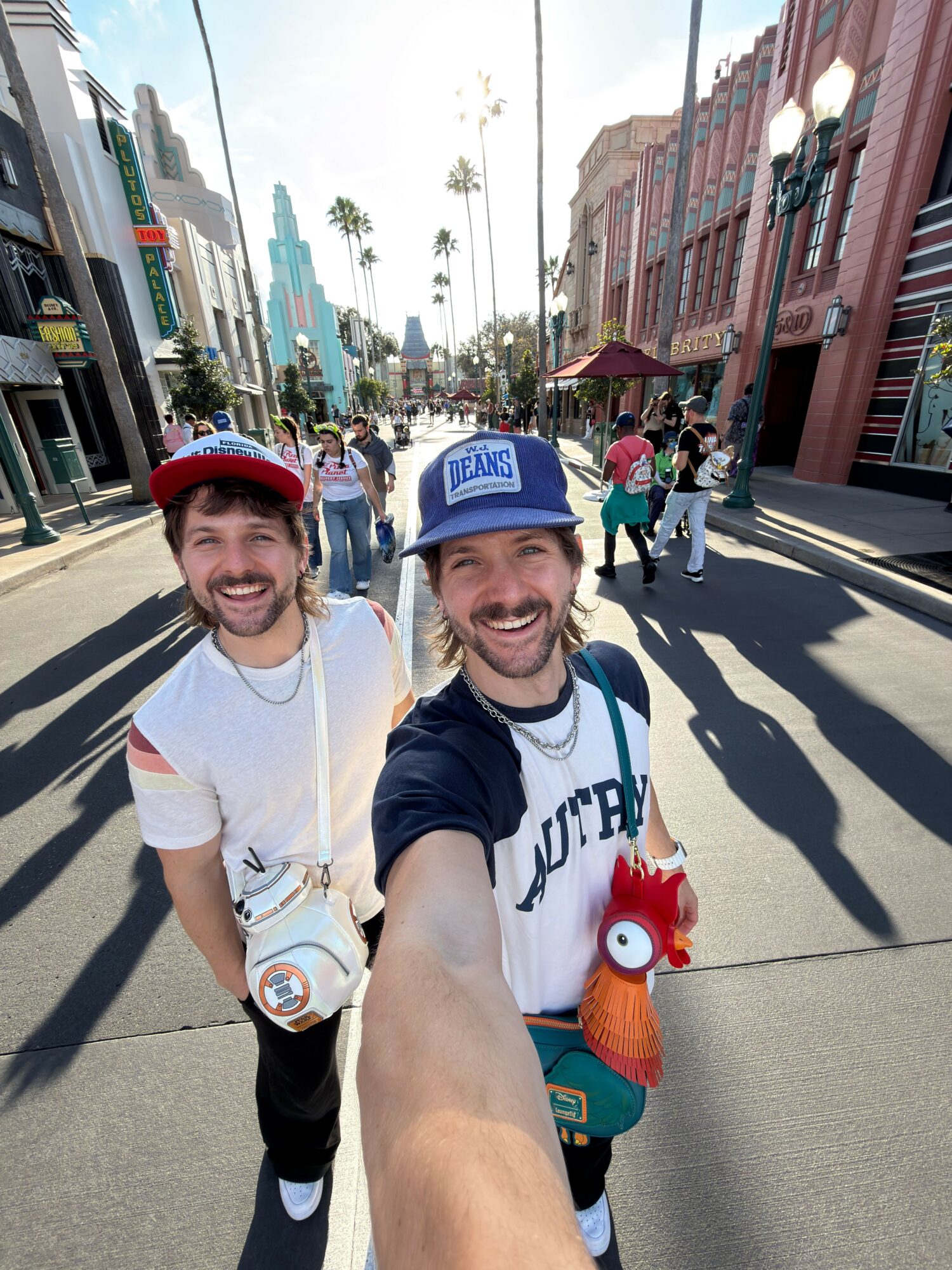 Two people taking a selfie on a busy street with buildings and pedestrians, one wearing a blue cap and the other a red cap.