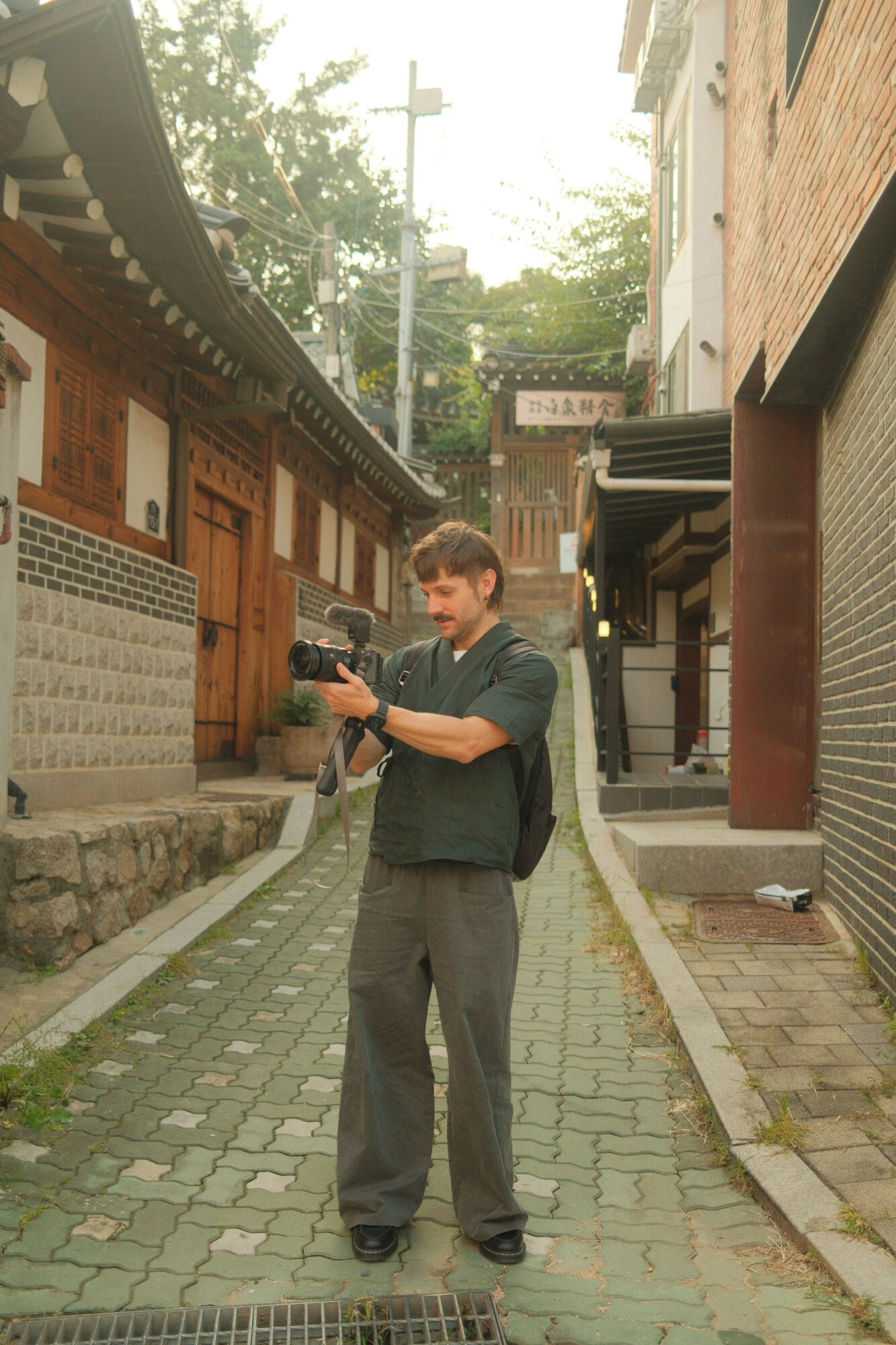 Man standing on a cobblestone street holding a camera, with buildings and trees in the background.
