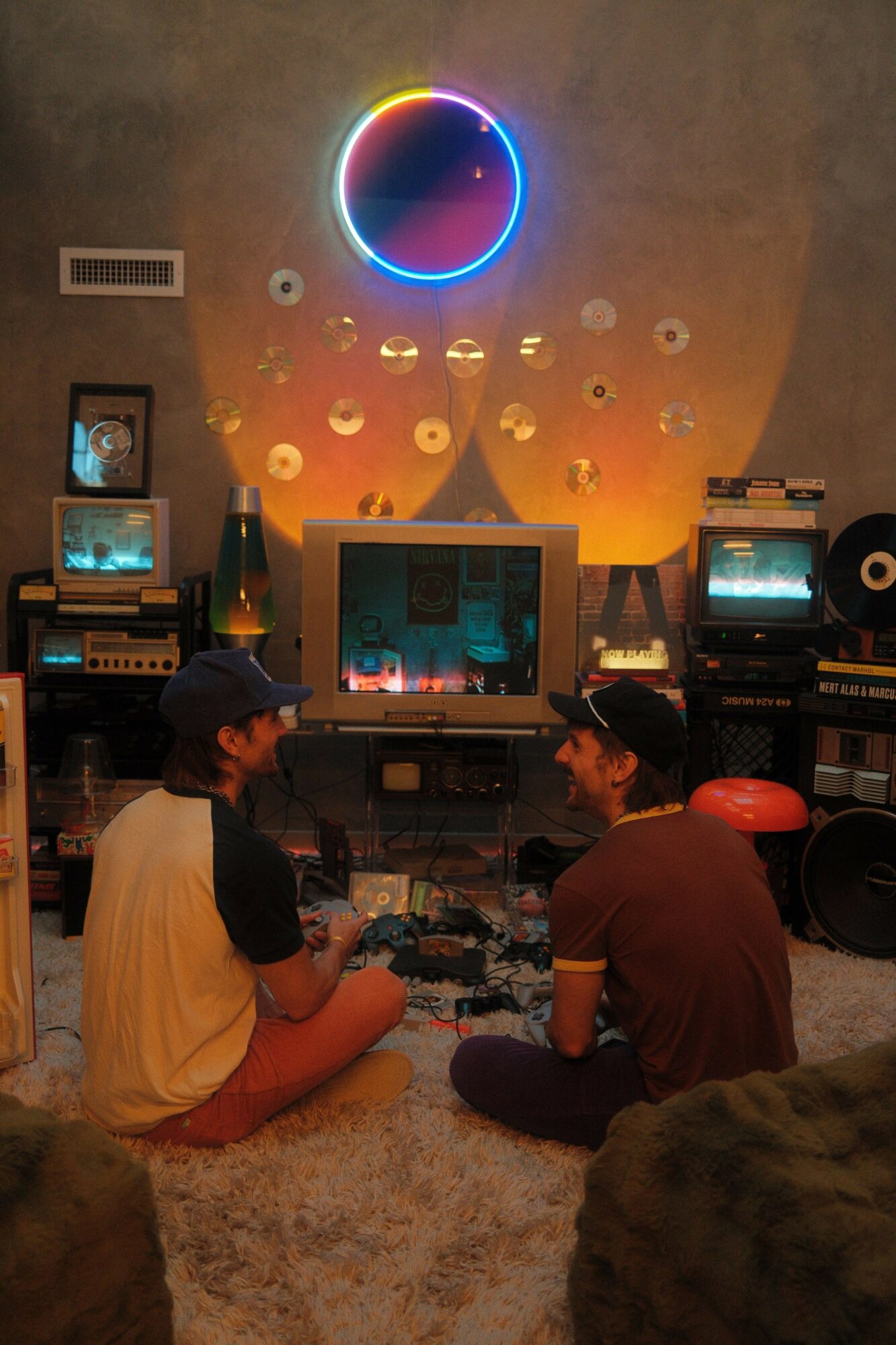 Two people sit on the floor watching a screen in a room with colorful circular lights on the wall.