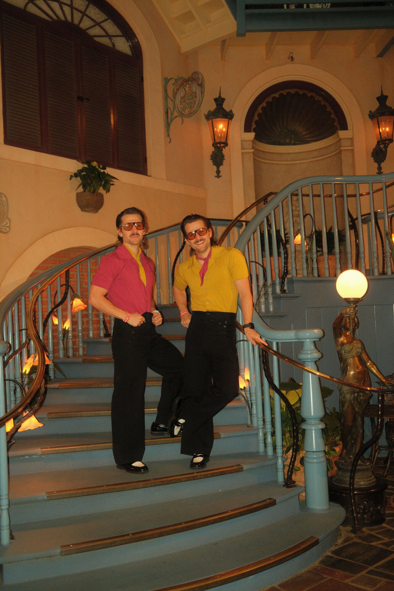 Two women standing on a staircase with ornate railing, smiling, in a decorated interior with plants and lighting fixtures.