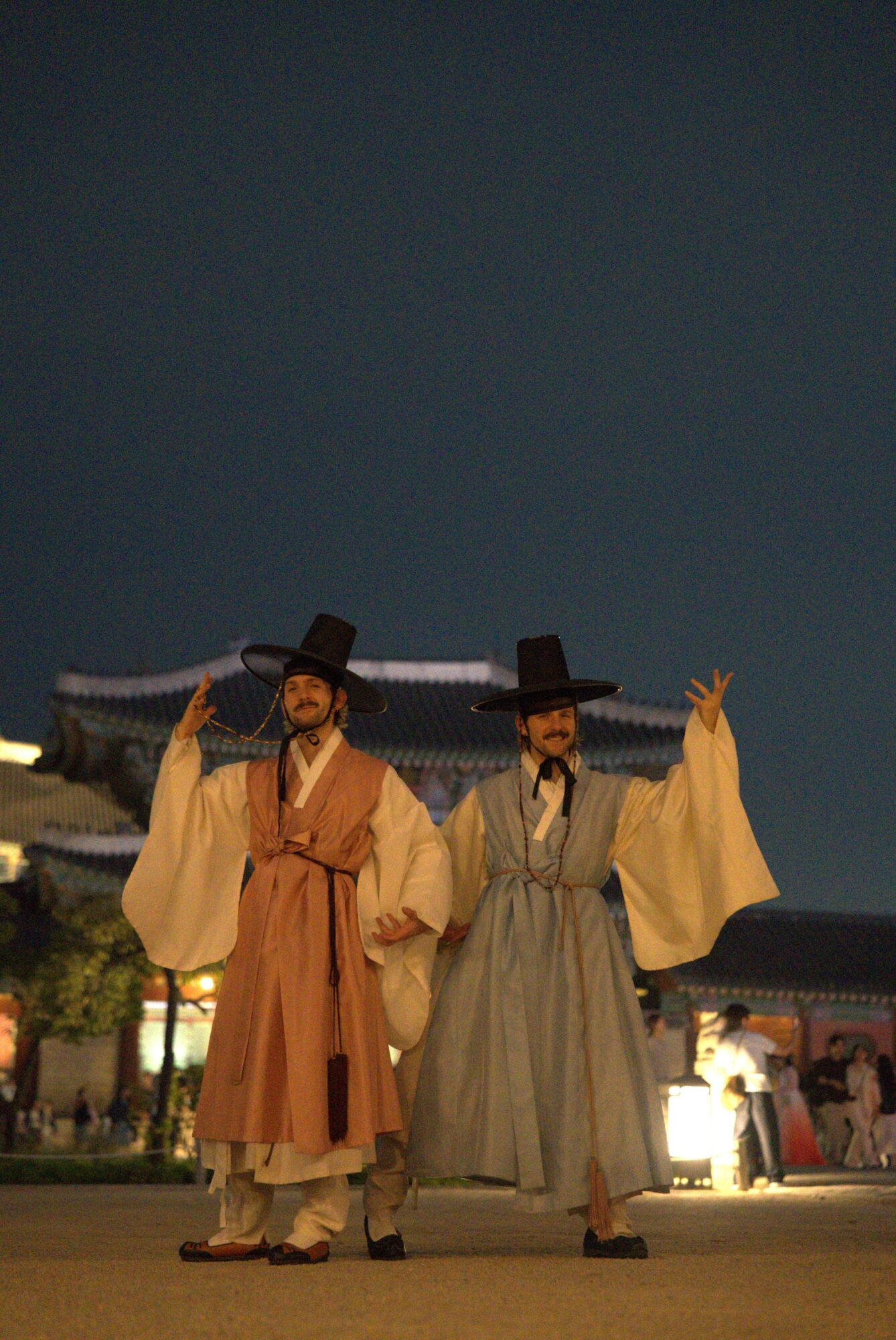 Two people in traditional costumes standing outdoors at night, with a building and other people in the background.