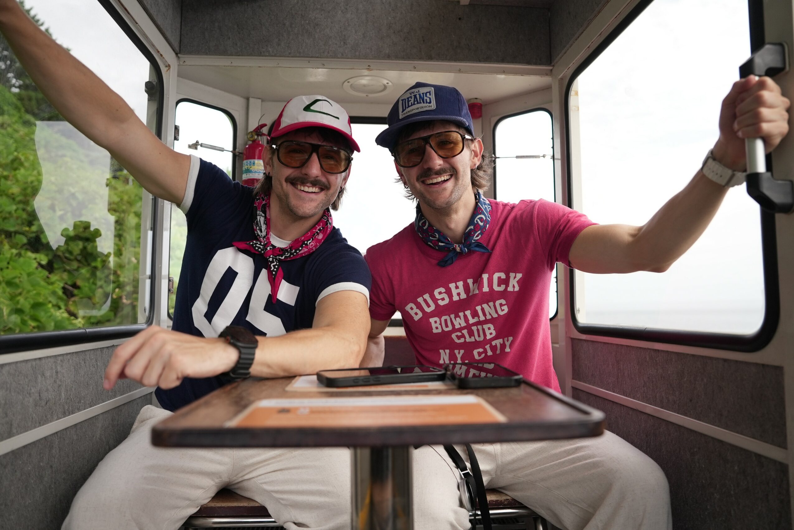 Two smiling men sitting inside a bus, holding onto handrails, wearing casual clothes and caps, with a window view outside.
