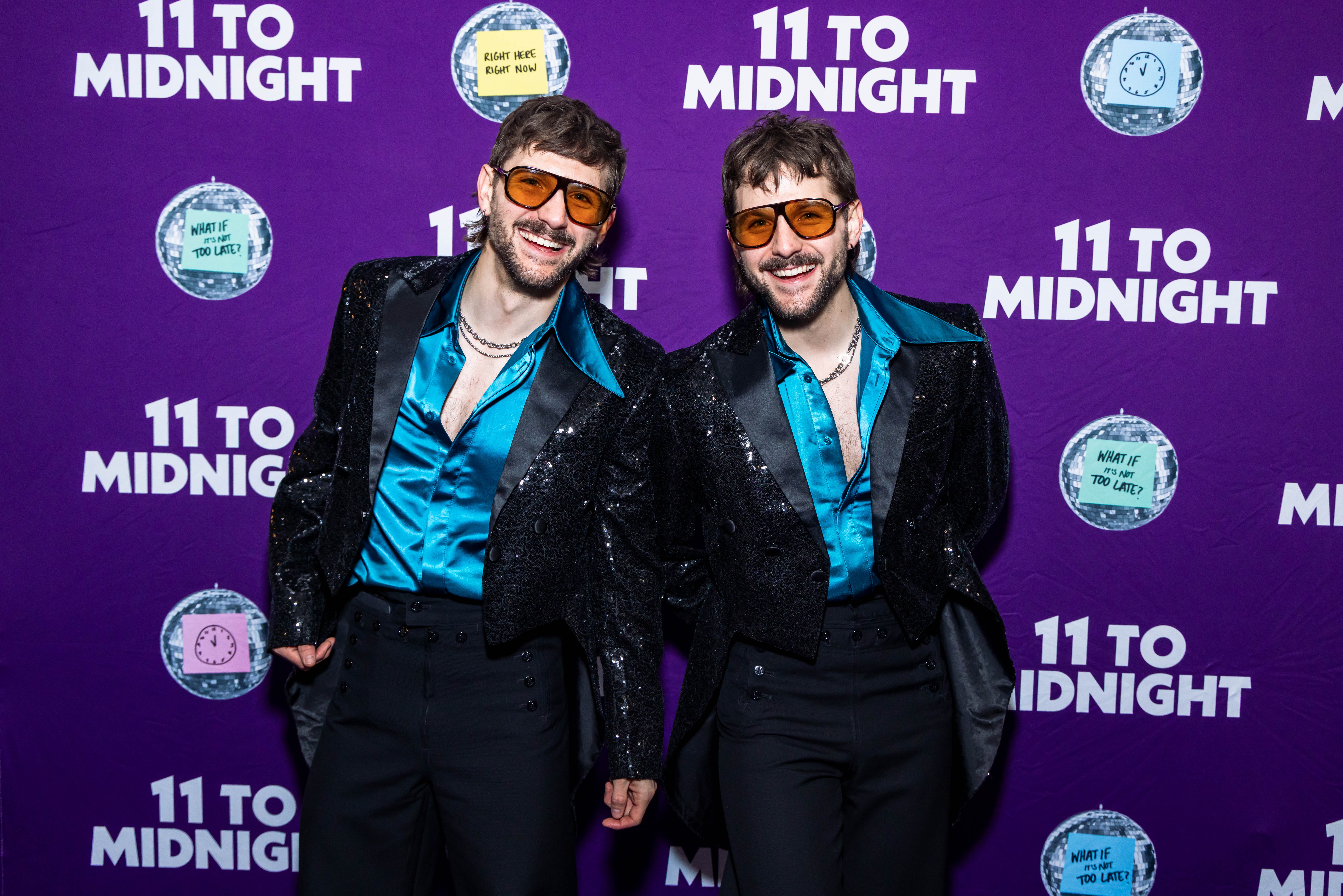 Two men in matching outfits with sunglasses, smiling, holding hands, standing in front of a purple backdrop with '11 to Midnight' text.
