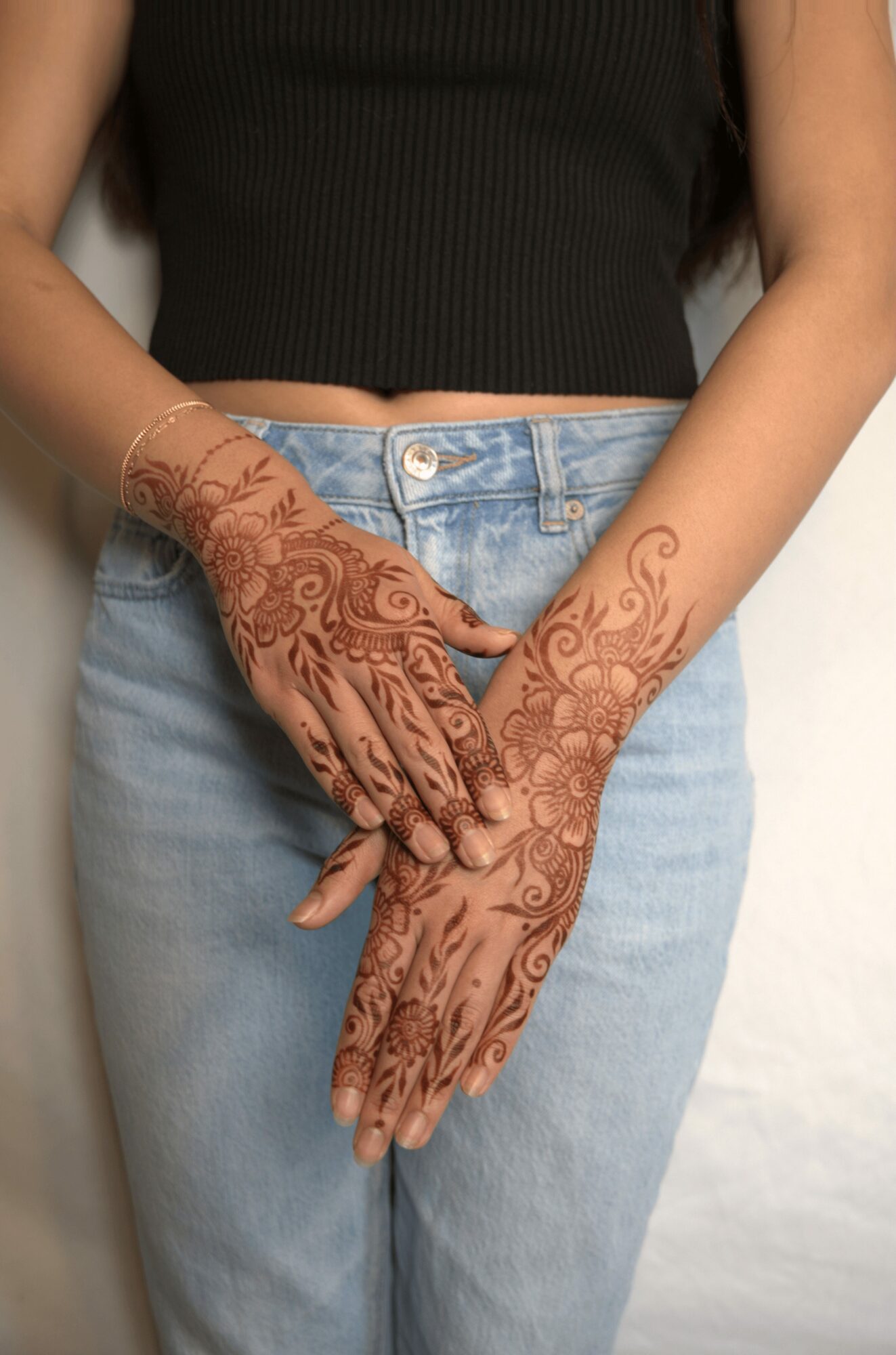 Person with hands decorated with henna, wearing a black top and light blue jeans, hands crossed in front.