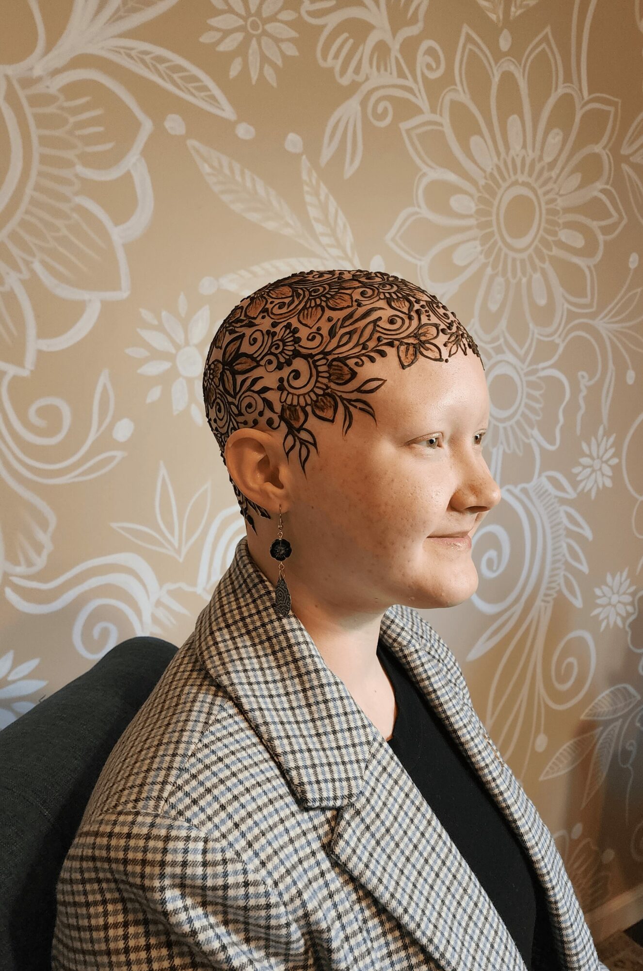 Woman with short hair and earrings, wearing a checkered blazer, sitting indoors with floral wallpaper background.