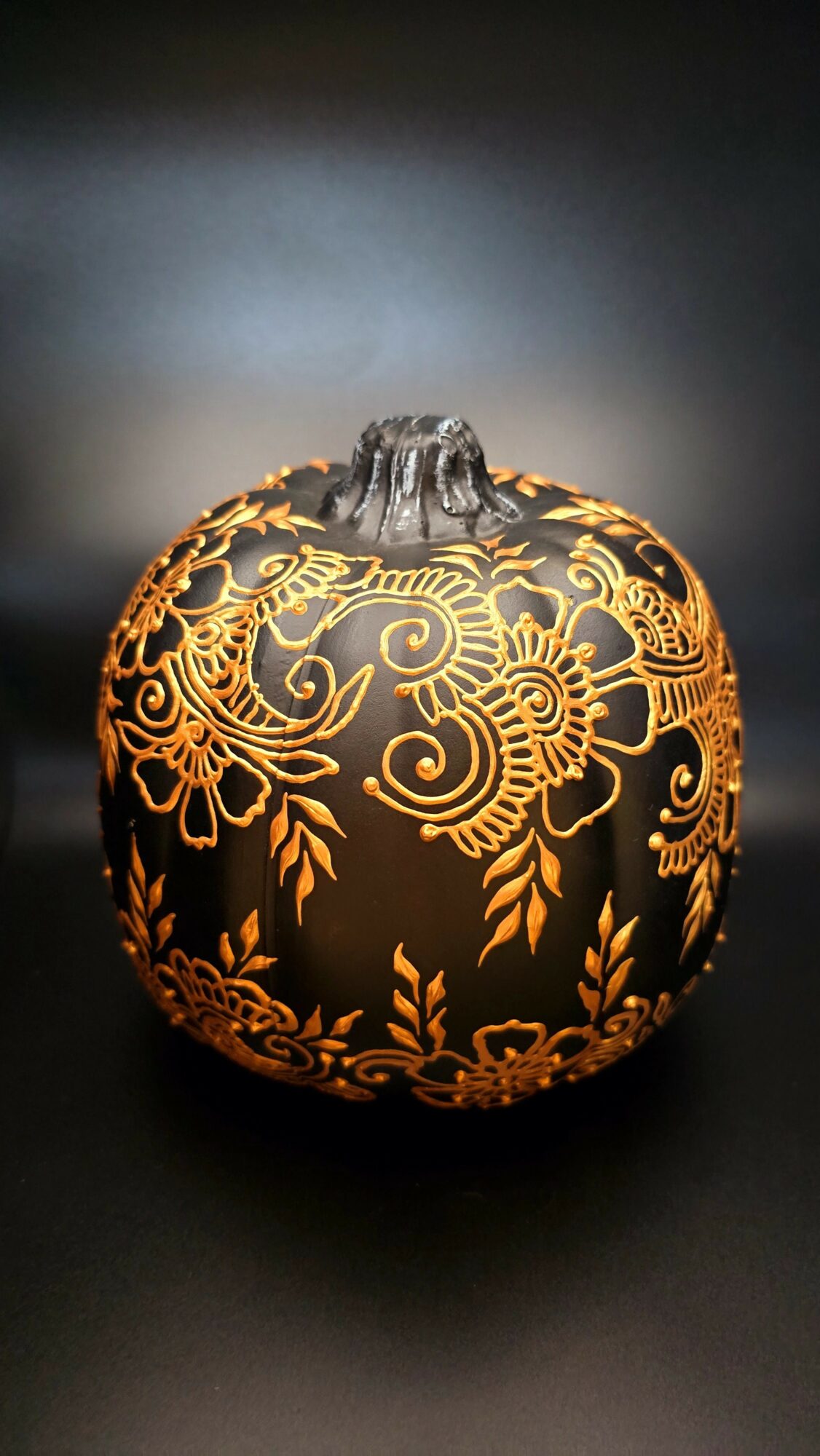 Decorative pumpkin with intricate gold floral patterns on a dark background, illuminated to highlight details.