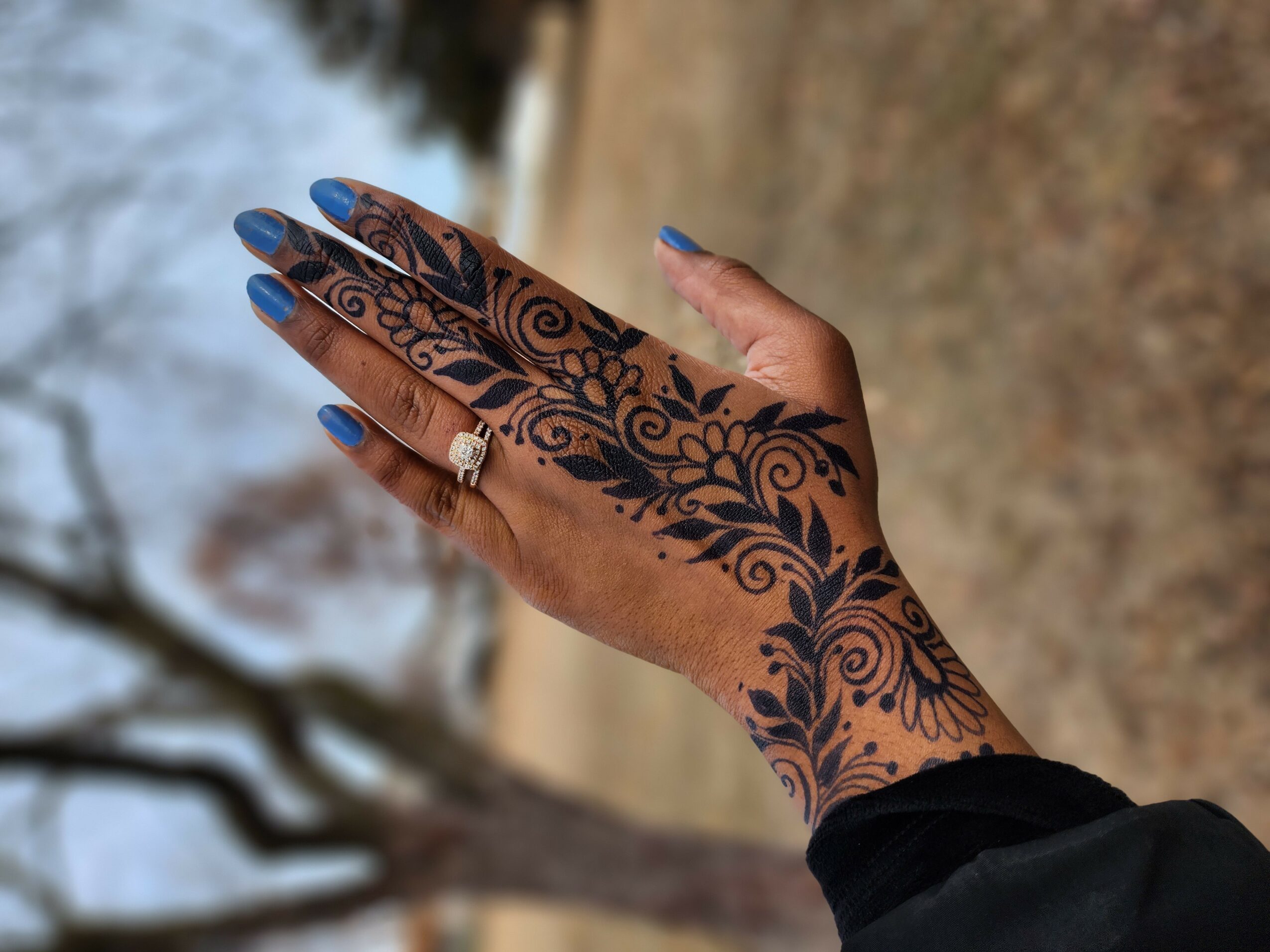 Hand with intricate henna design, blue nail polish, and a ring, outdoors with trees and ground in background.