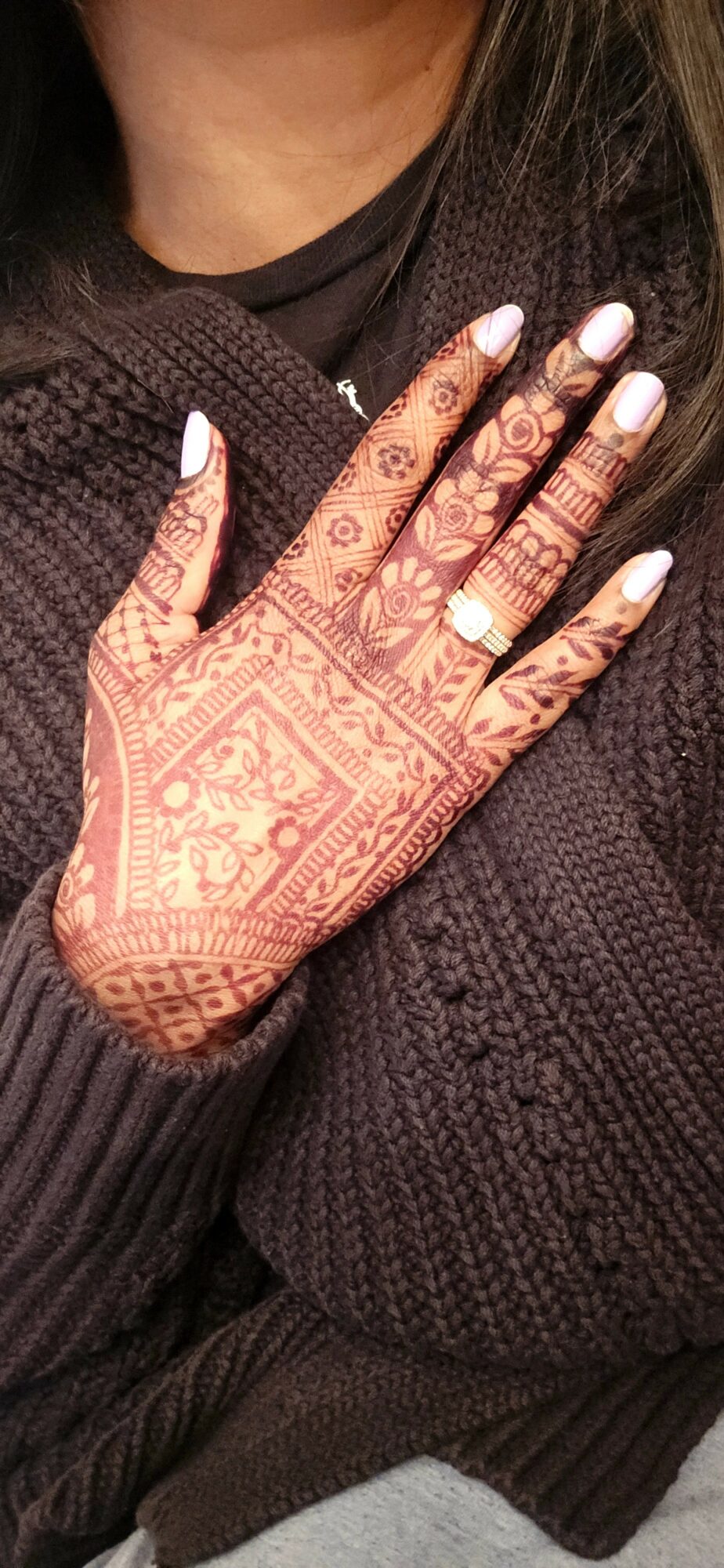Person's hand with intricate henna designs, white nail polish, and a ring, resting on a dark sweater.