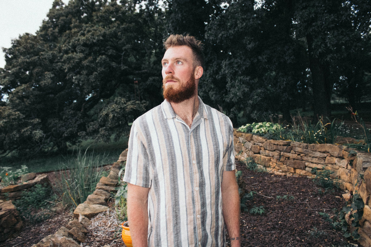 Man with a beard and short hair wearing a striped short-sleeve shirt outdoors, looking to the side.