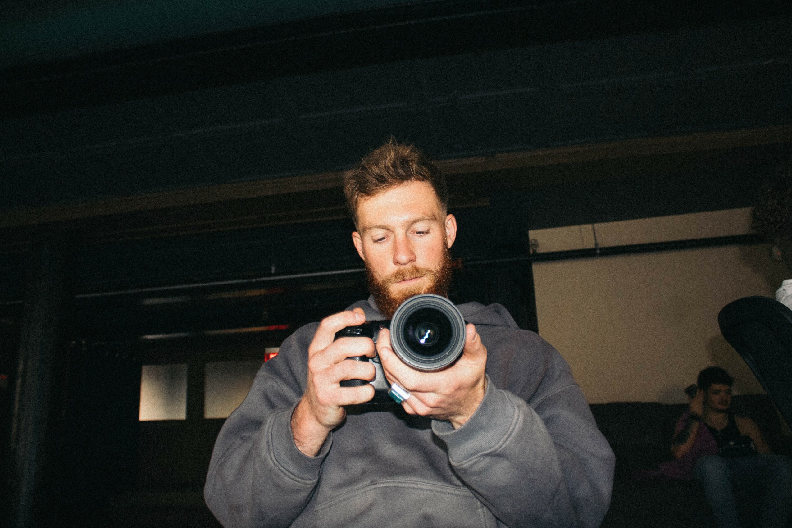 Man with beard taking a photo indoors, holding a camera, with a dark ceiling and people in the background.
