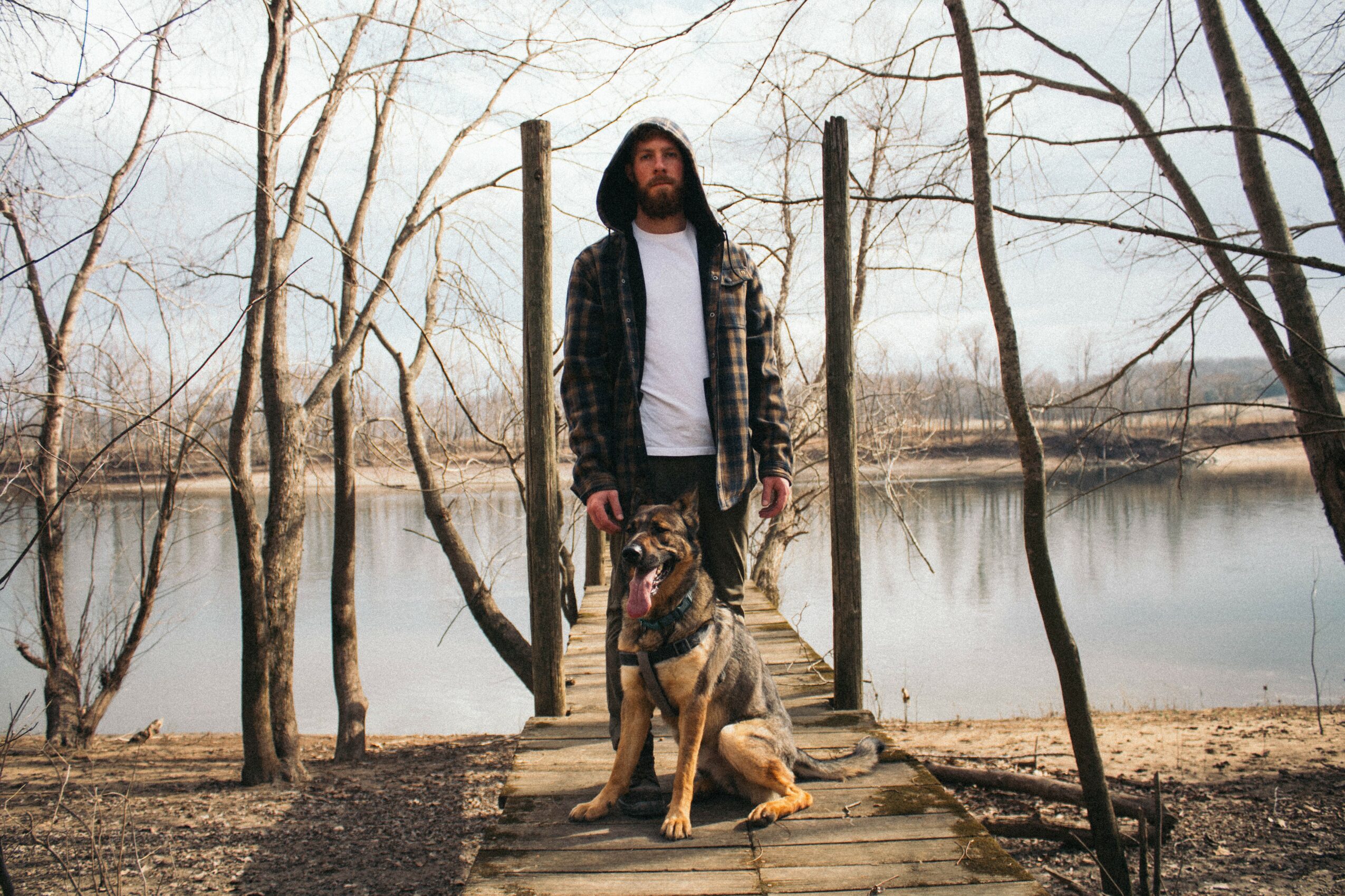Man with long hair and beard standing on a wooden dock by a lake, with a dog sitting beside him, surrounded by leafless trees.