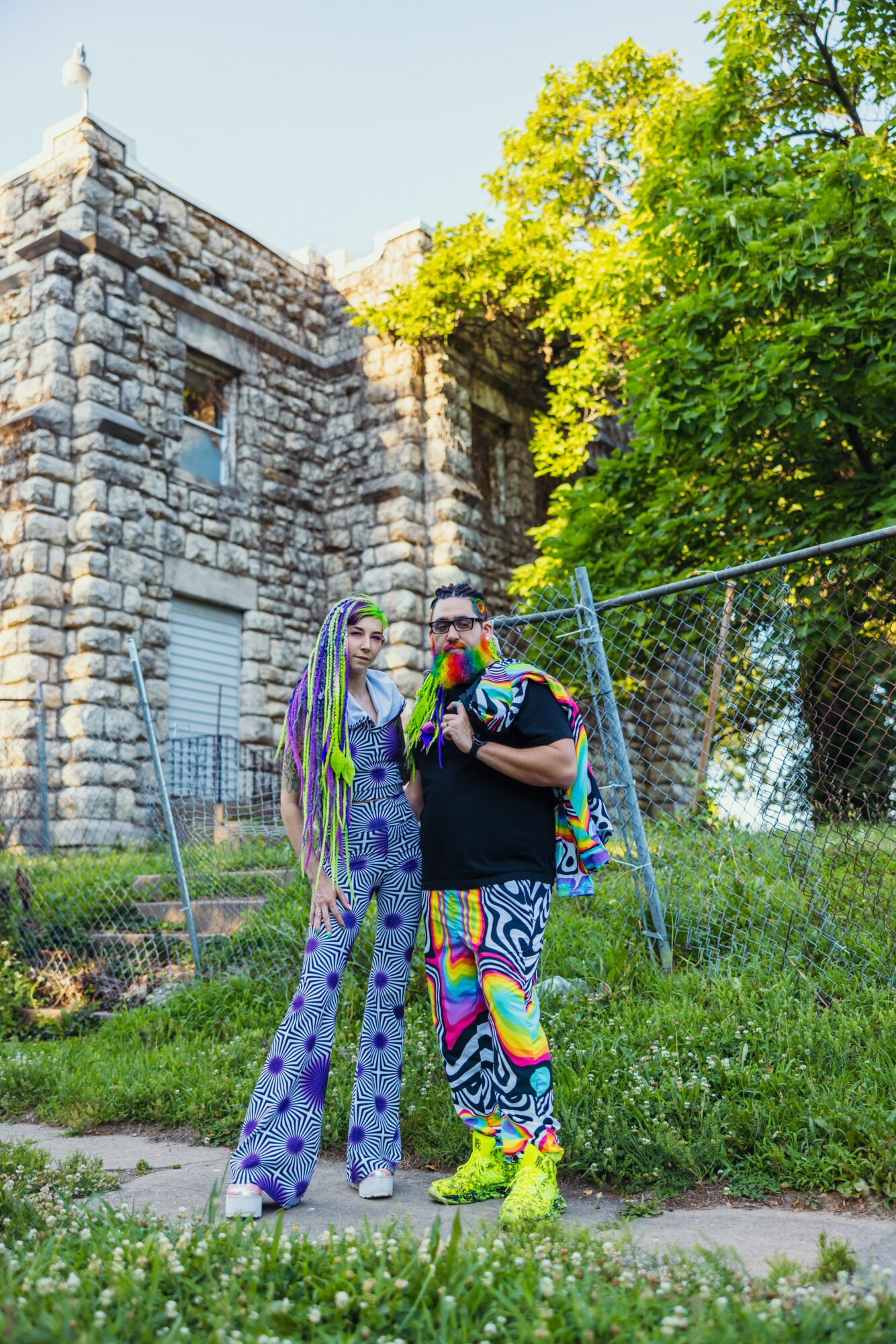 Two people stand outdoors near a stone building and green trees, one wearing colorful patterned pants and the other in a long dress.