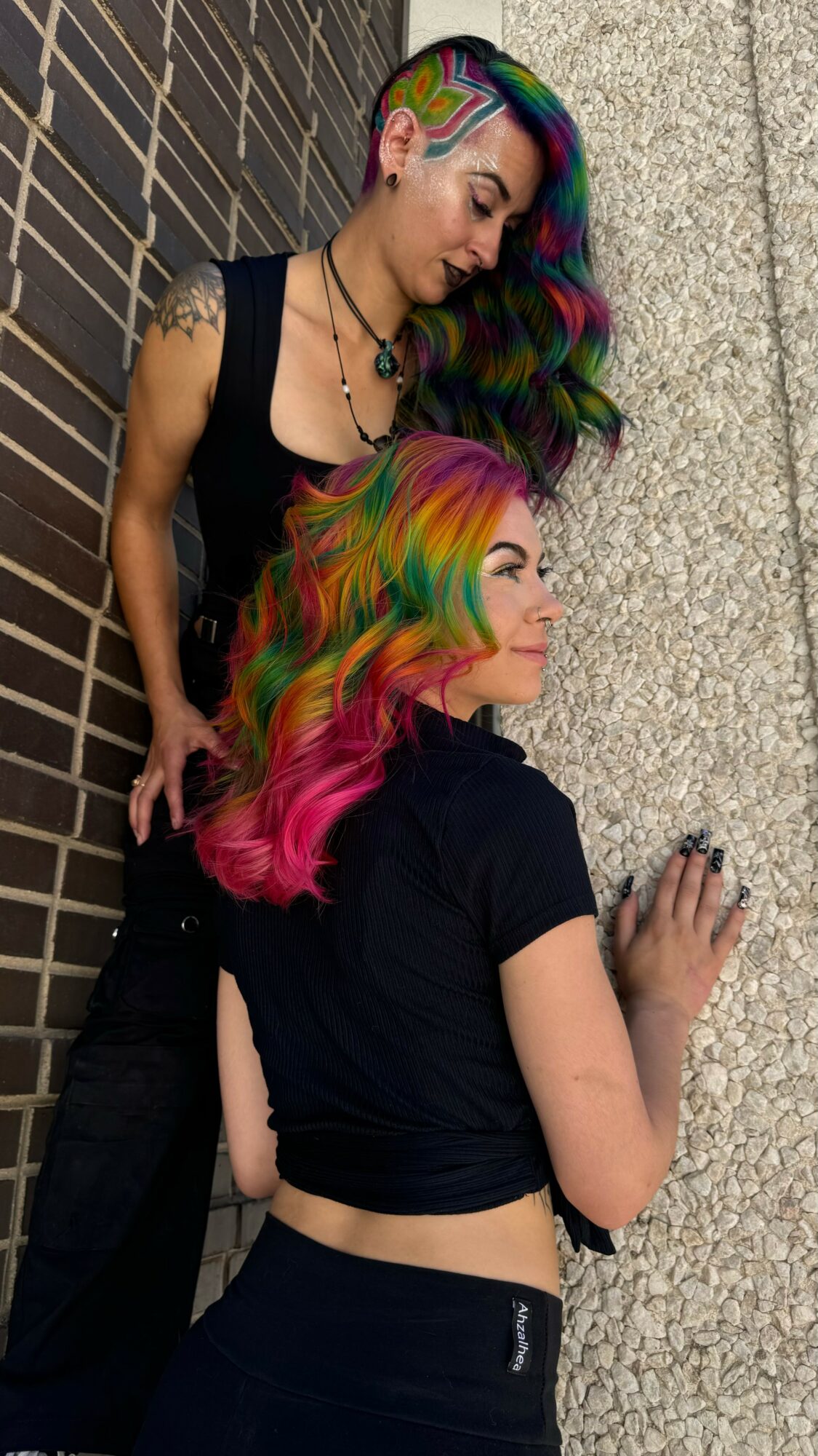 Two women with colorful hair leaning against a brick and concrete wall, one looking down, the other looking to the side.