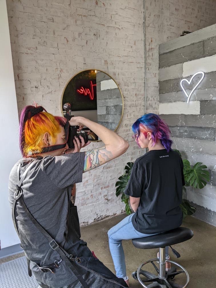 Person with colorful hair taking a photo of another person with rainbow-colored hair sitting on a stool, in a room with white brick wall.