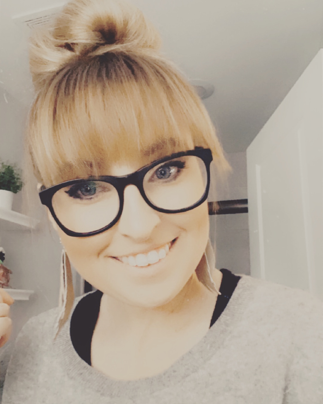 Young woman with blonde hair in a bun, wearing glasses, smiling, in a room with white walls and shelves.