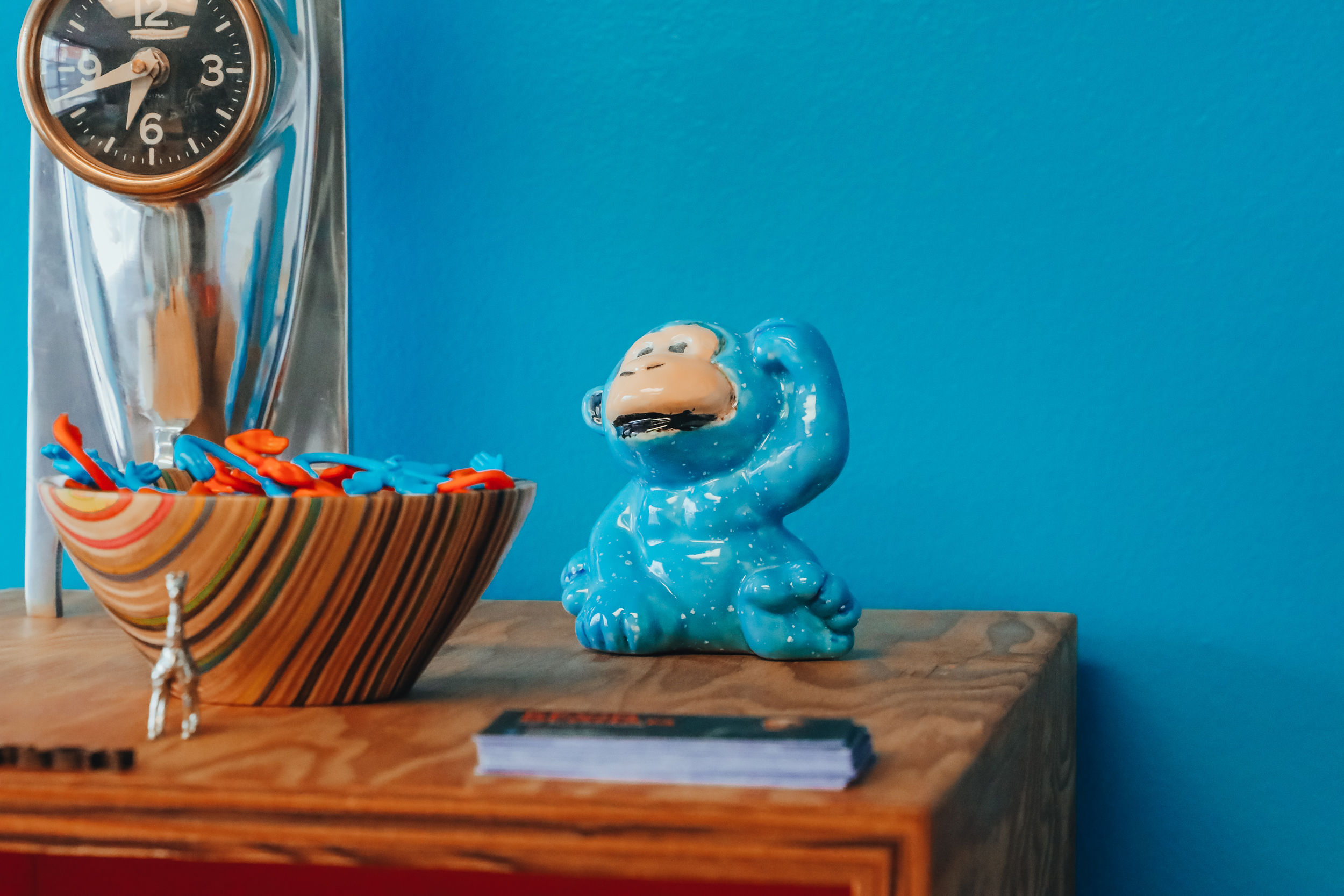 Blue dinosaur figurine on wooden surface with a bowl and clock in background, against a blue wall.