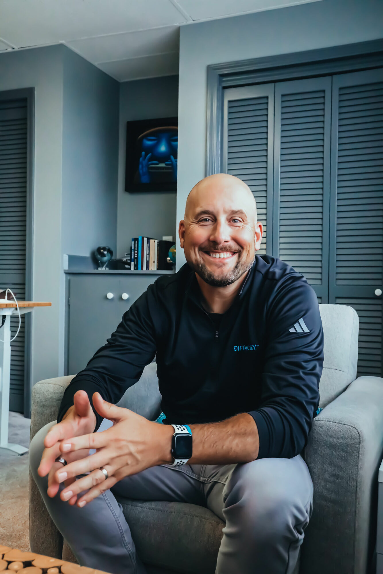 Man with a beard smiling, sitting on a gray couch in a room with blue walls and closed shutters.