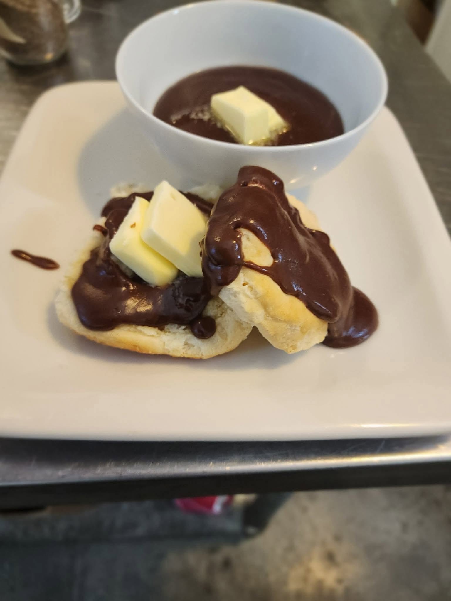 Banana slices topped with chocolate sauce and butter pieces on a white plate, with a bowl of chocolate sauce in the background.