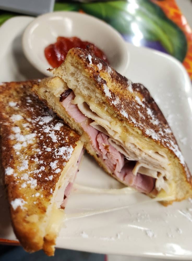 Sandwich with ham and cheese, cut in half, on a white plate with a small dish of ketchup in the background.