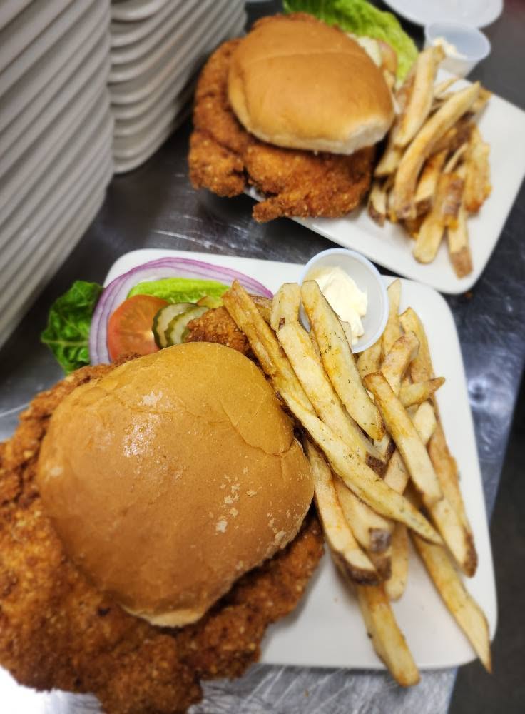 Two plates with fried chicken sandwiches, French fries, and pickles, on a dark surface. One sandwich has a bun, fried chicken, and lettuce.
