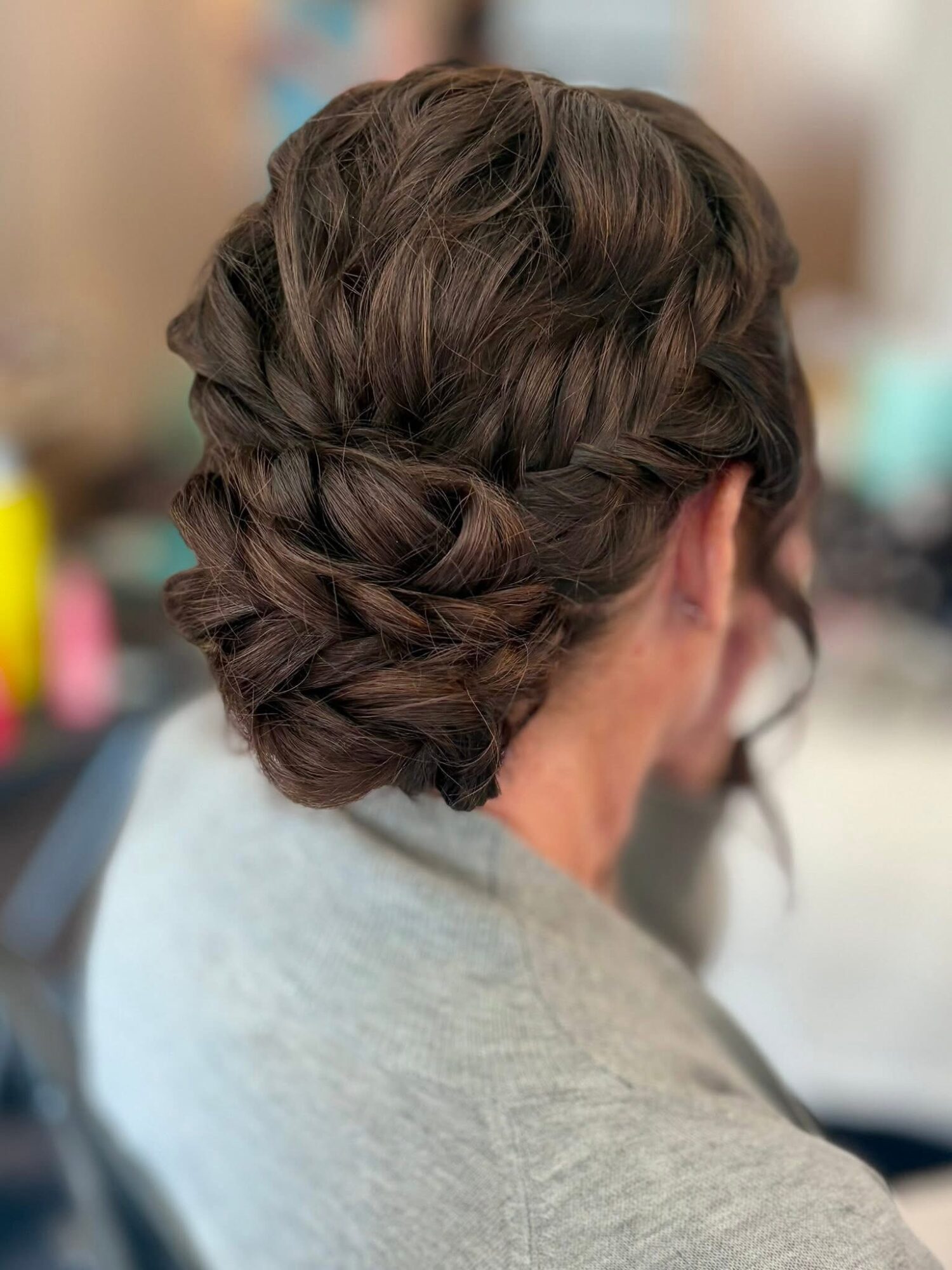 Back view of a woman with styled brown hair in an updo, wearing a light gray top.