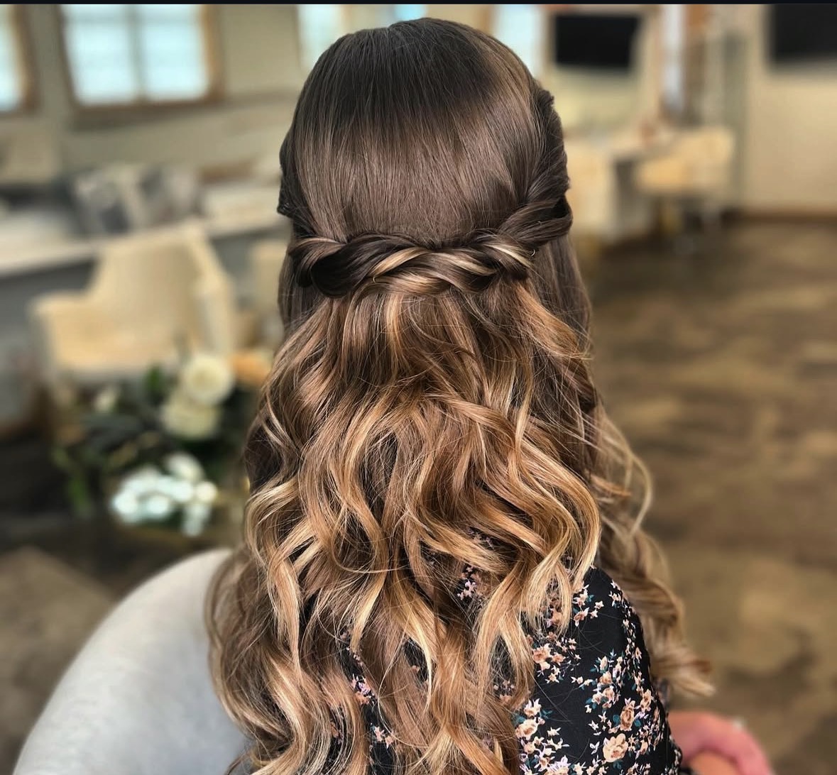 Back view of a woman with long, wavy hair styled with a twisted half-updo, in a cozy indoor setting.