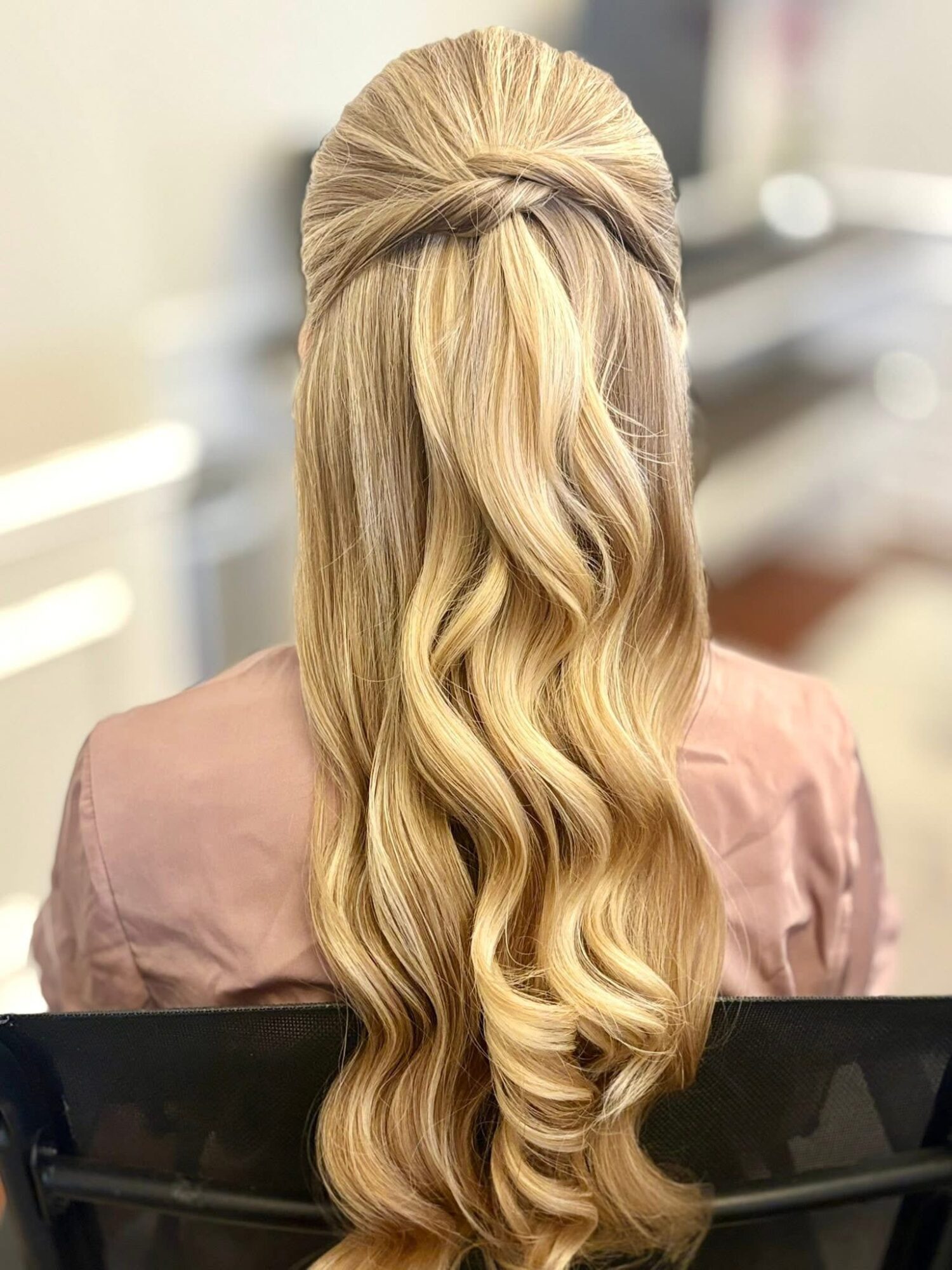 Back view of a woman with long, wavy blonde hair styled with a half-up twist, sitting in a salon chair.