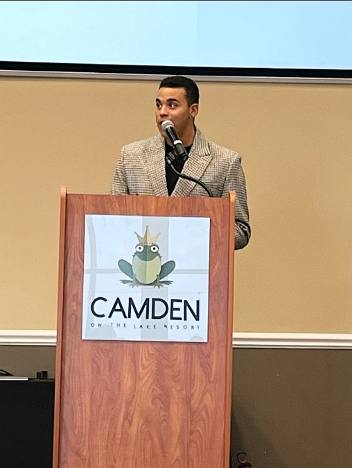 Man speaking at a podium with a Camden logo, in front of a beige wall and a projection screen.
