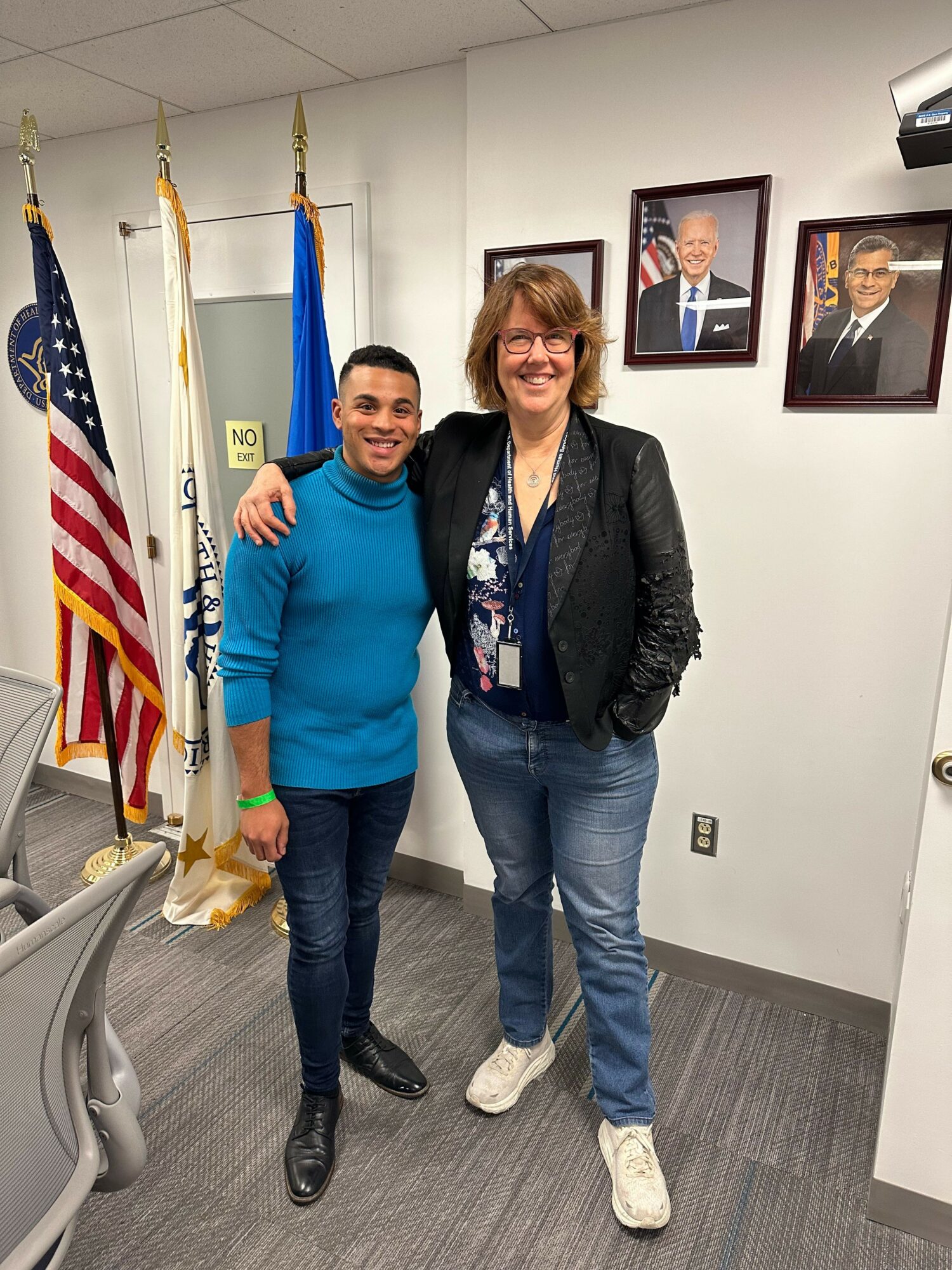 Two women standing indoors, one in a black jacket and jeans, the other in a blue sweater and jeans, smiling and giving a thumbs-up. Flags and framed pictures are in the background.