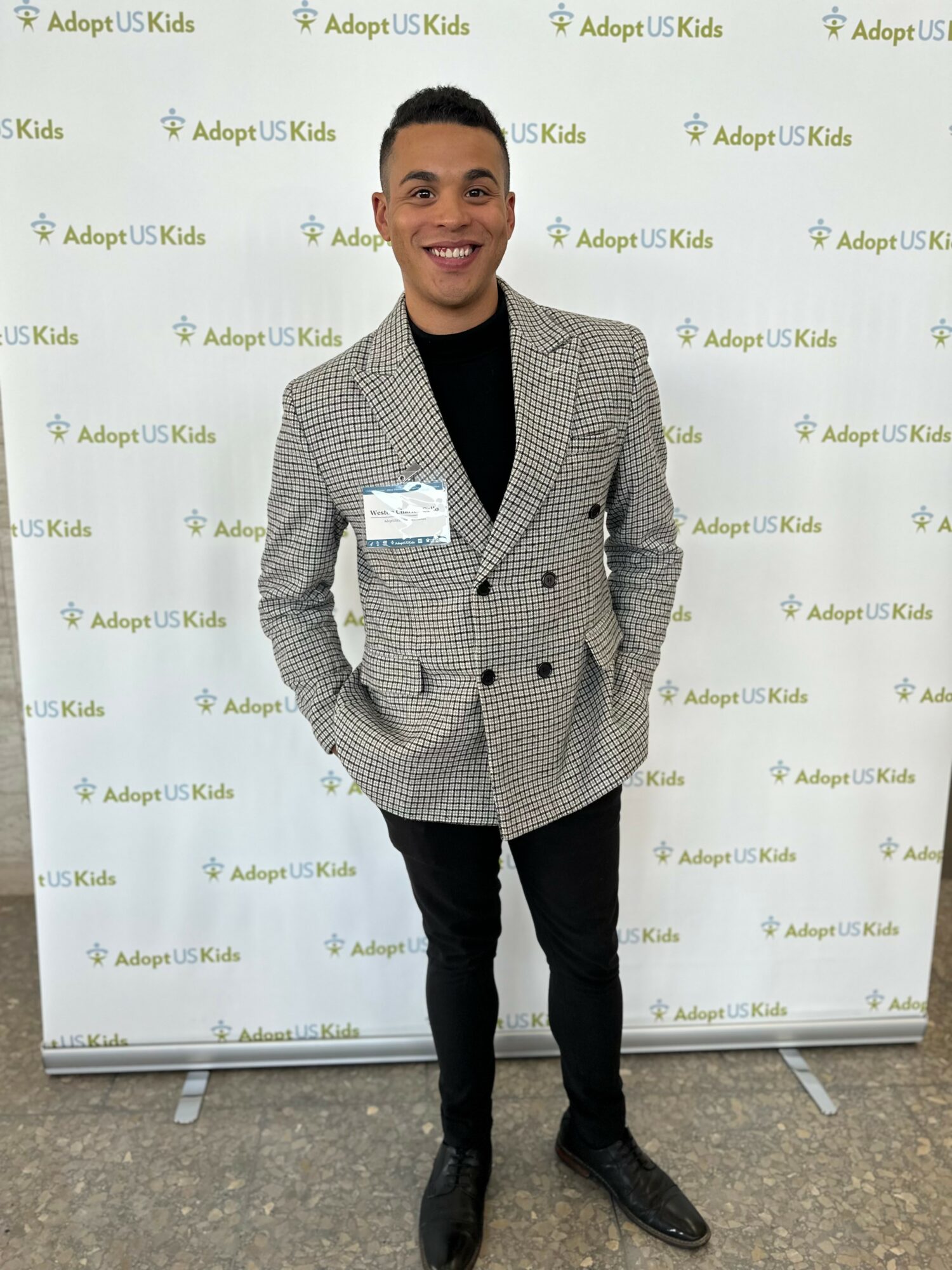 Person standing in front of a backdrop with 'Adopt US Kids' logo, smiling, wearing a gray checkered blazer and black pants.