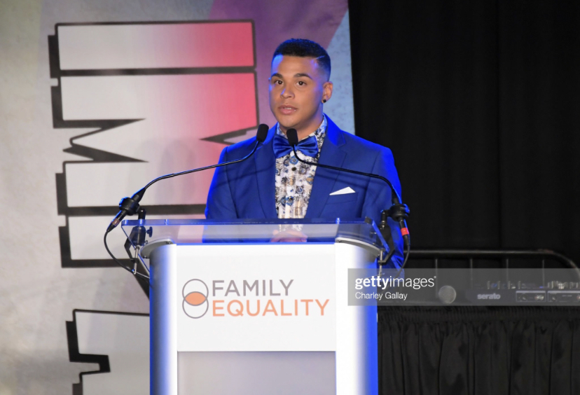 Young man in a blue suit speaking at a podium with a sign reading 'Family Equality' during an event.
