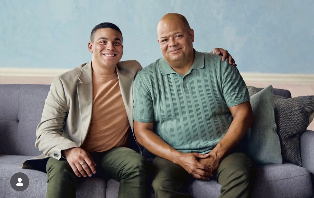 Two men sitting on a gray couch, smiling, with a light blue wall behind them.
