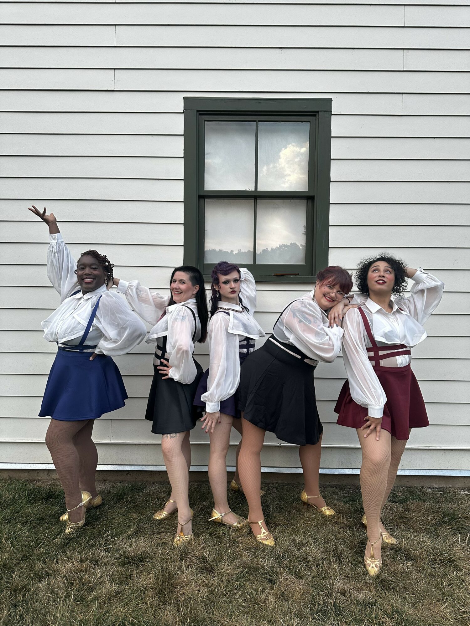 Five women in vintage-style outfits pose in front of a white house with a window, standing on grass.