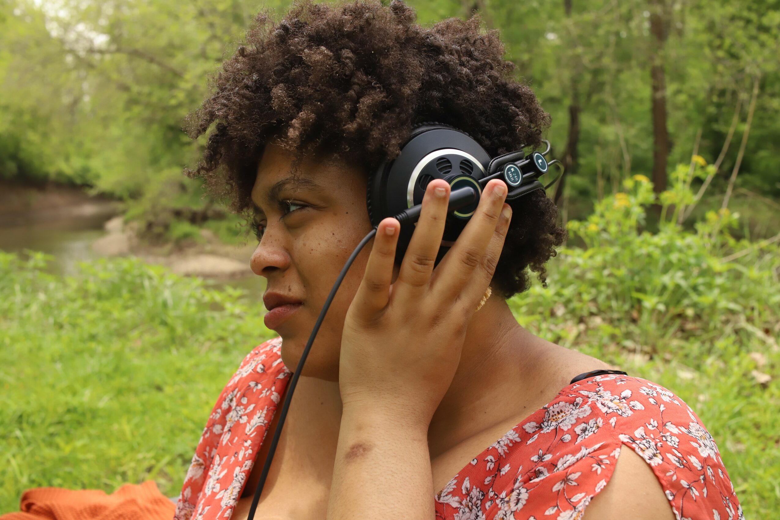 Young woman with curly hair wearing a red floral top, listening to headphones outdoors in a green natural setting.
