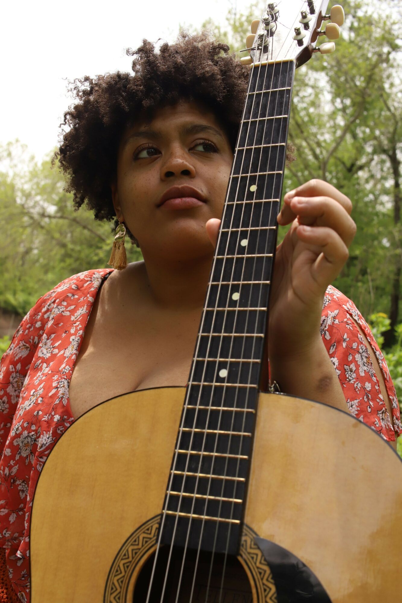 Person with curly hair holding an acoustic guitar outdoors, wearing a red patterned top, trees in background.