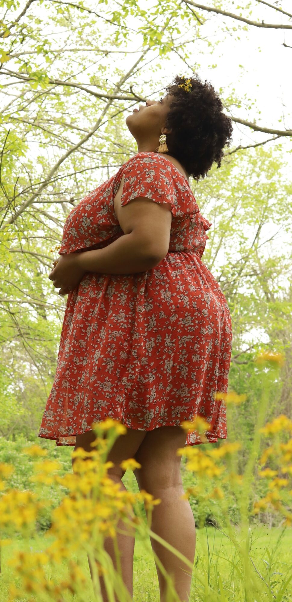 Woman in red floral dress standing outdoors among yellow flowers and green trees, looking upward.