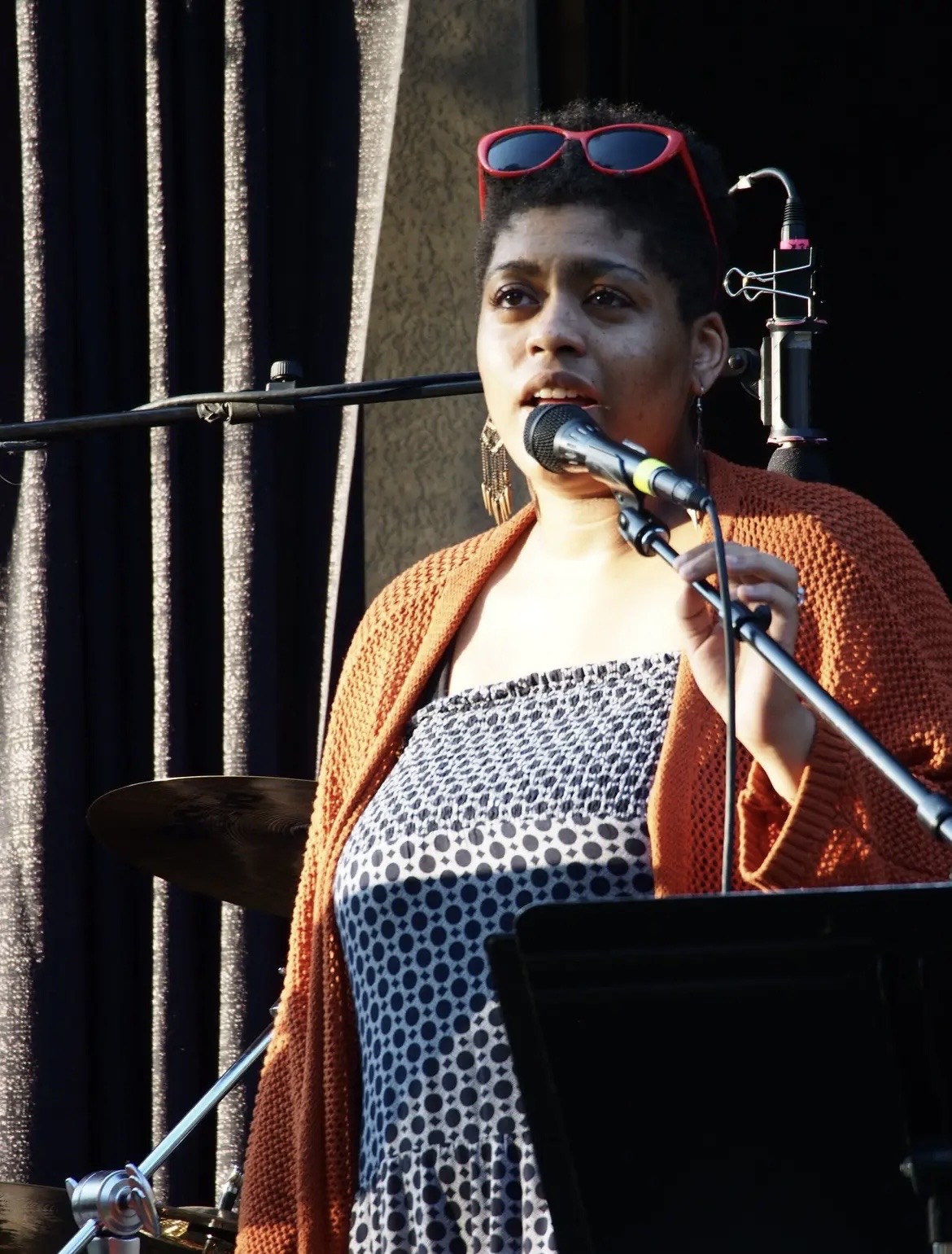 Woman singing into a microphone with a dark curtain background, wearing sunglasses on her head and an orange cardigan.