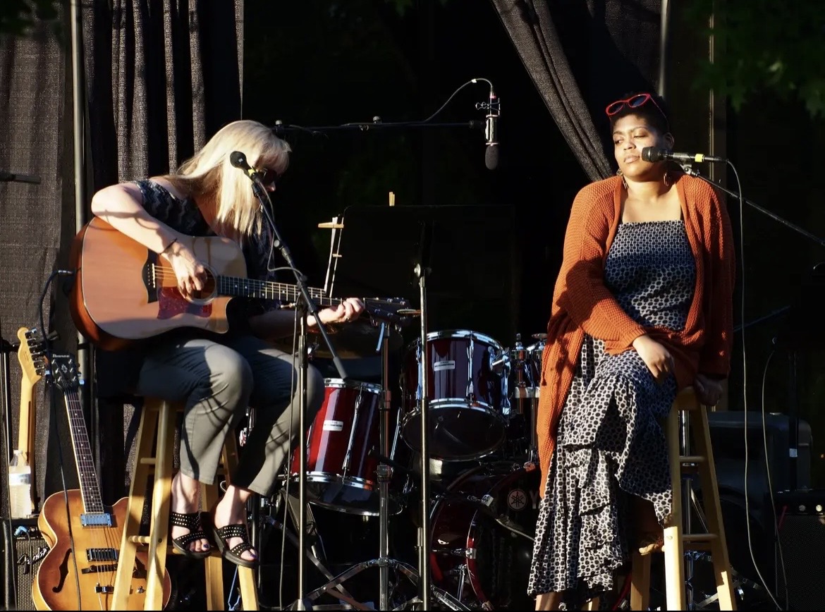 Two women perform on stage with musical instruments, one playing guitar and the other singing, in a dimly lit setting.