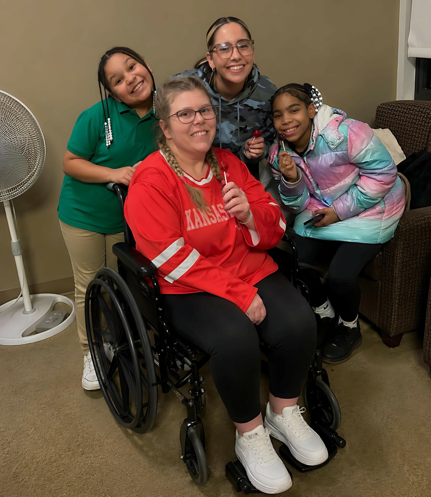 Four women, one in a wheelchair, smiling and posing together indoors, with a fan and a chair in the background.