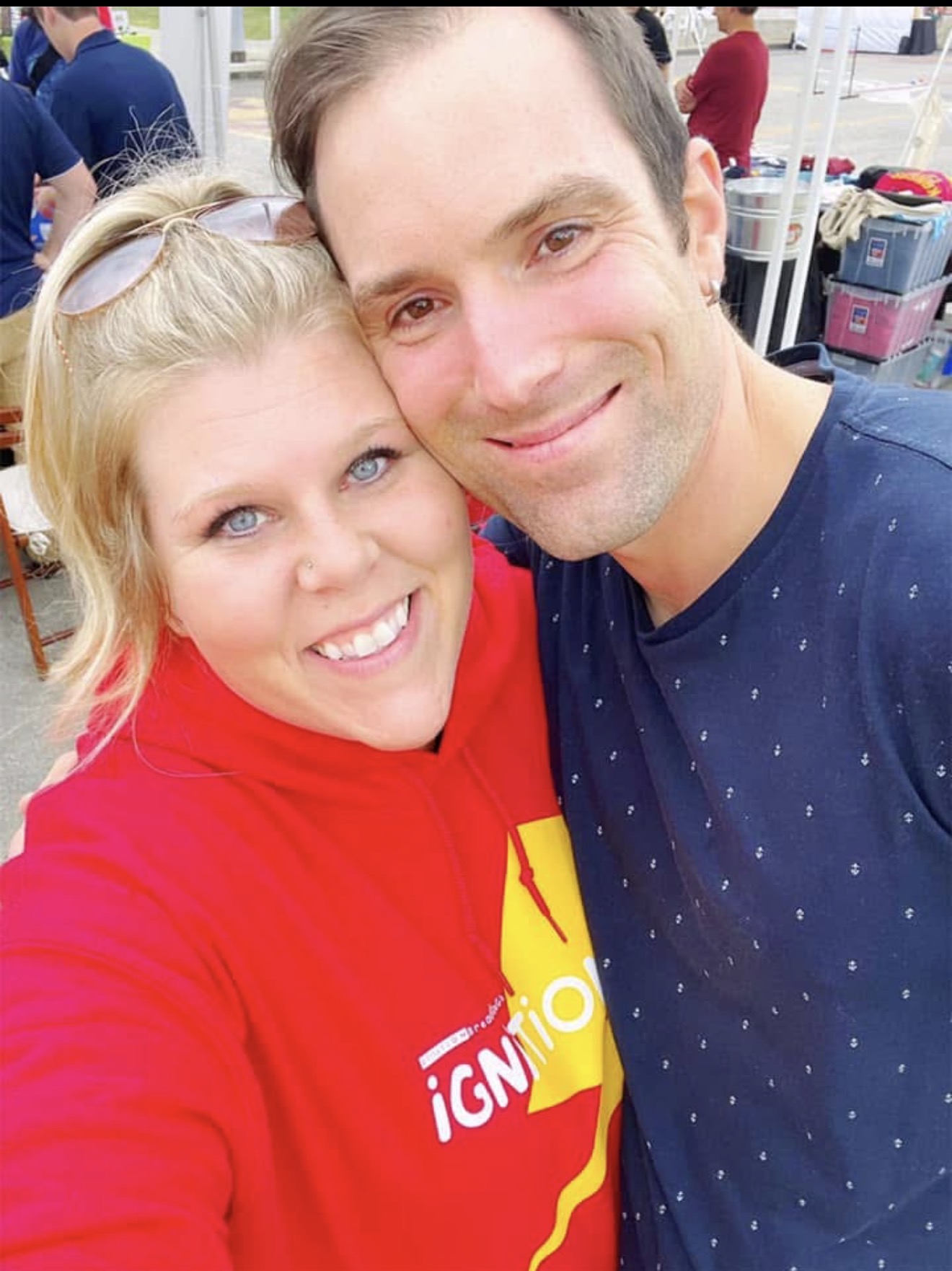 Two people smiling closely, woman in red hoodie and man in dark blue shirt, outdoors with blurred background