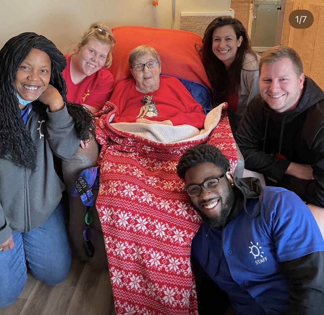 Group of five people gathered around an elderly woman in a hospital bed, smiling, with a red blanket and festive decorations.
