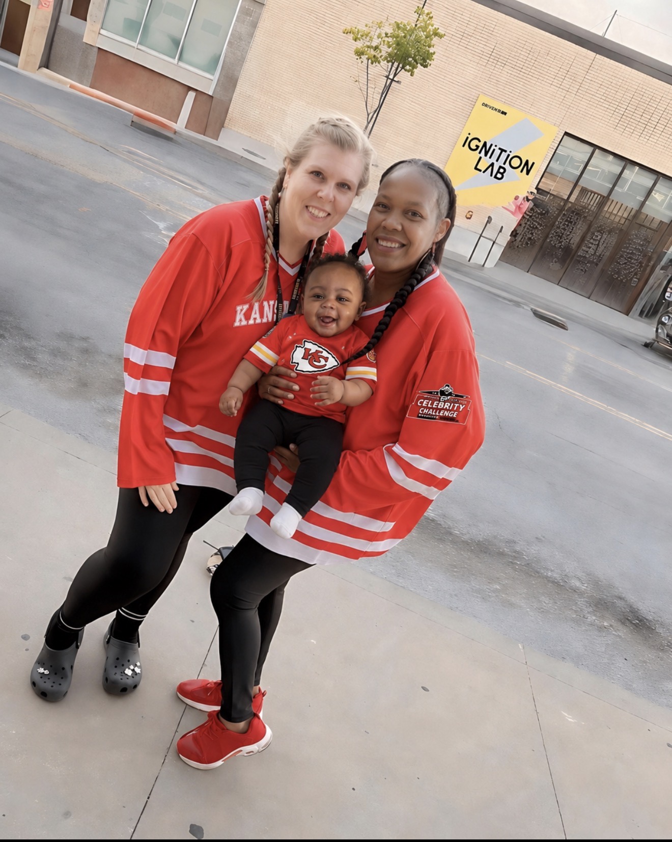 Two women and a baby standing outdoors on a sidewalk, smiling, with a building and a yellow sign in the background.