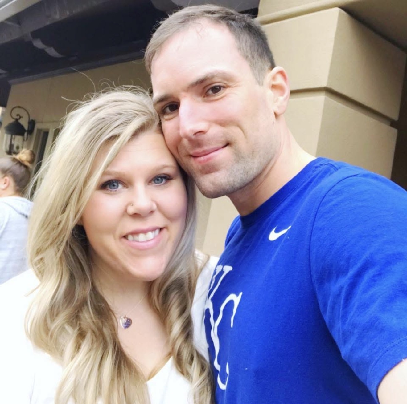 A man and woman smiling, close together, in an outdoor setting with a building in the background.