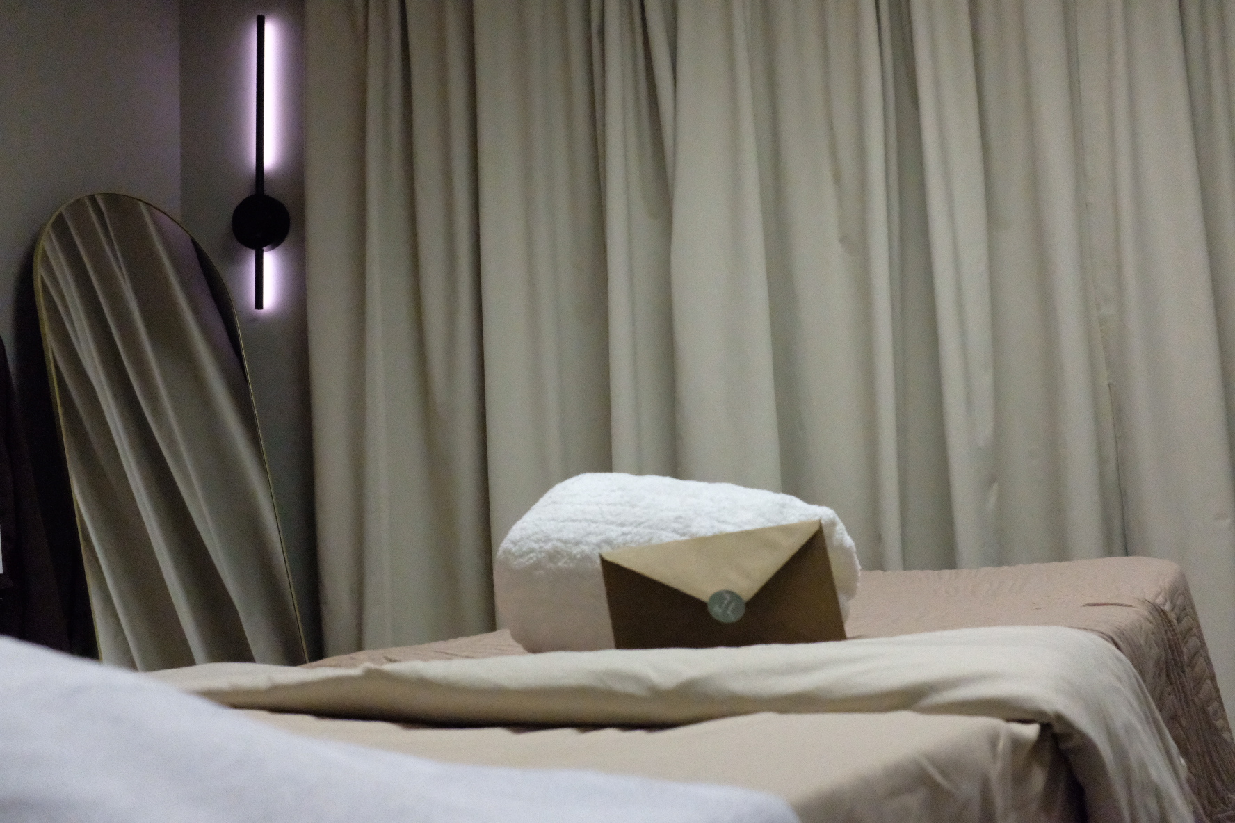 A massage table with a pillow and blanket in a room with curtains and a mirror, dim lighting.