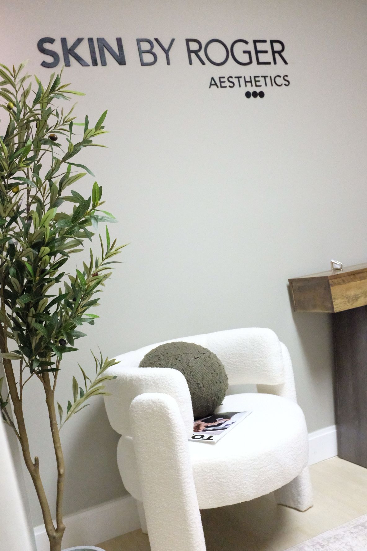 A white chair with a round cushion, a plant, and a wall with the text 'SKIN BY ROGER AESTHETICS'.