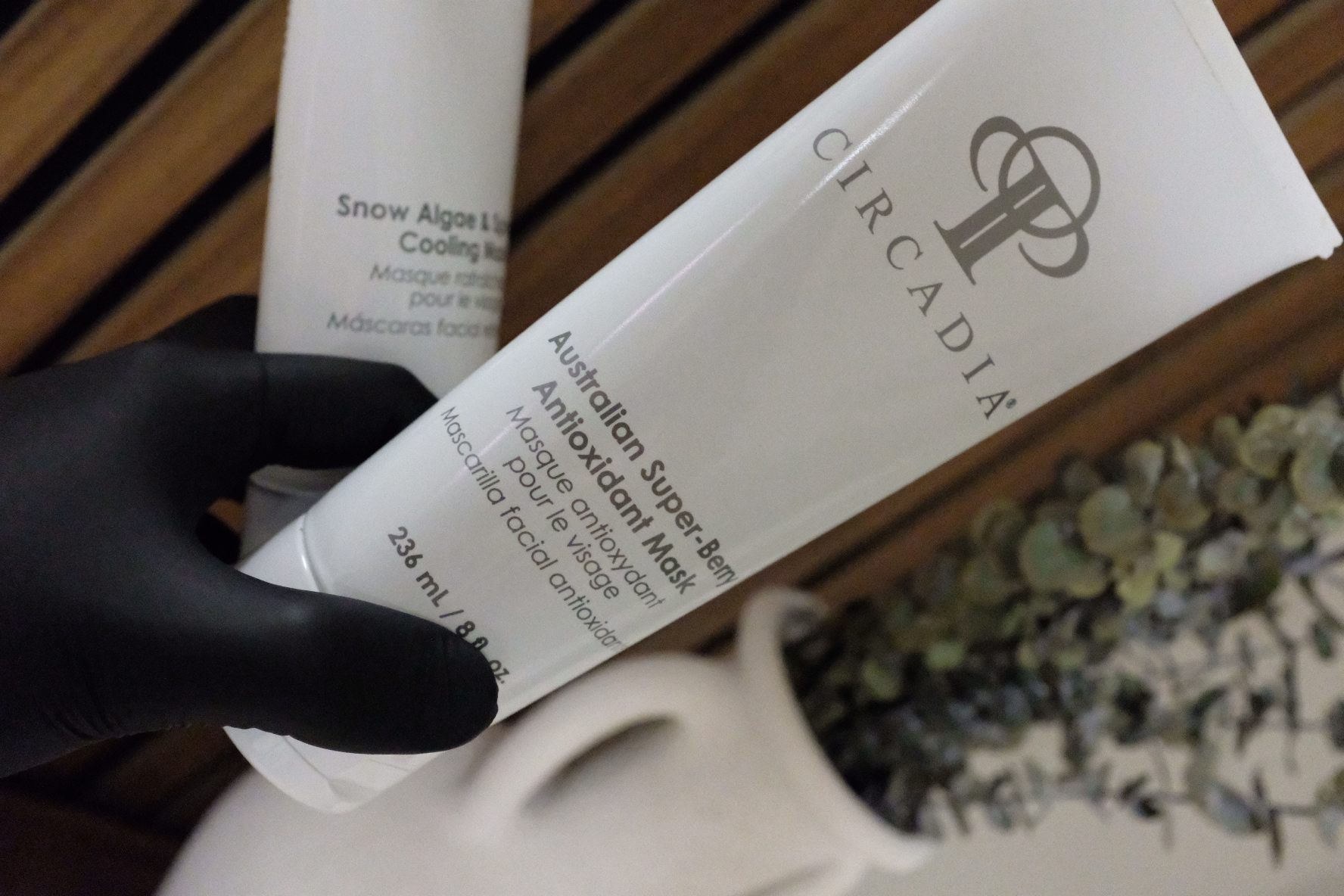 Person wearing black glove holding a white tube of skincare product near a white vase with flowers, on a wooden surface.