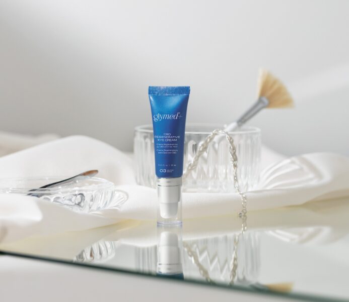 Blue skincare tube on a reflective surface with a glass container and a fan in the background.